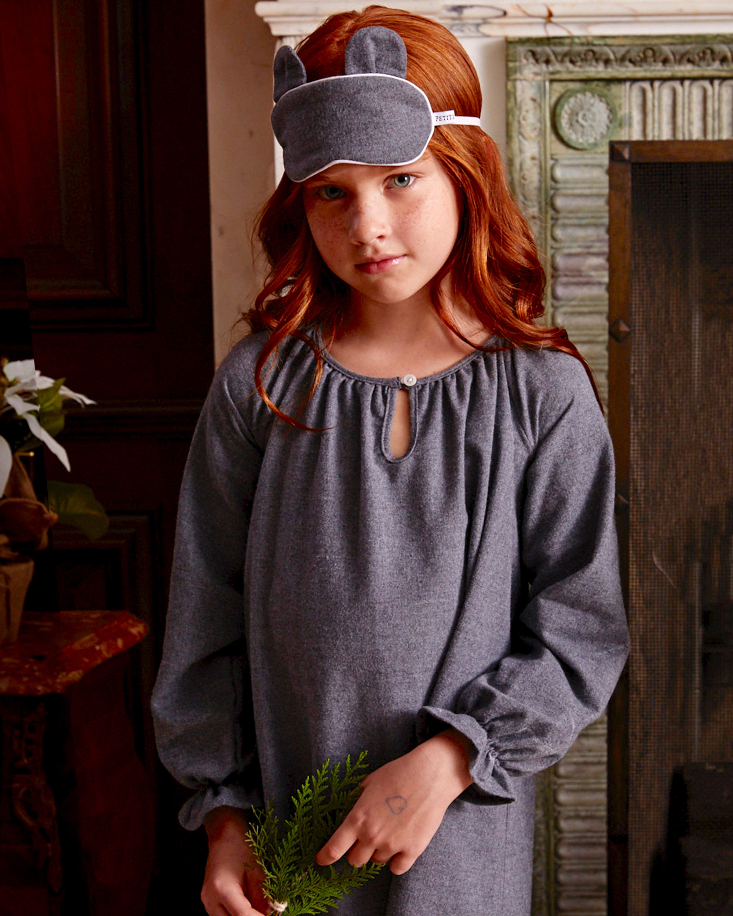 A young red-haired girl stands indoors wearing the Petite Plume Girls Flannel Delphine Nightgown in Grey, paired with a matching sleep mask featuring bear ears. She cradles a small green plant in front of an elegant decorative fireplace.