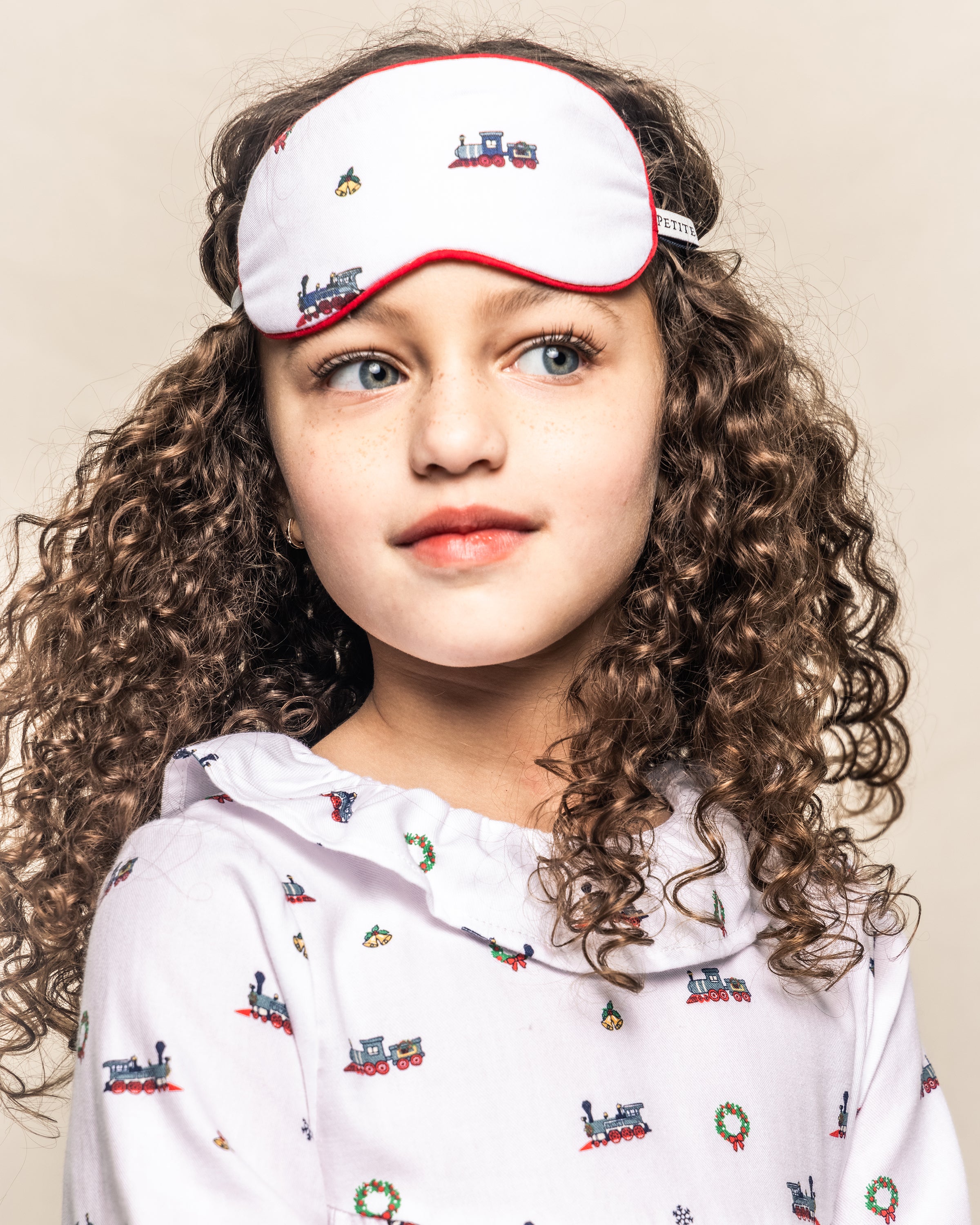 A young girl with curly hair wears the Girls Twill Scarlett Nightgown in Arctic Express by Petite Plume, featuring train designs and a matching white sleep mask. She gazes slightly to the side against a neutral background, embodying luxurious sleepwear comfort.