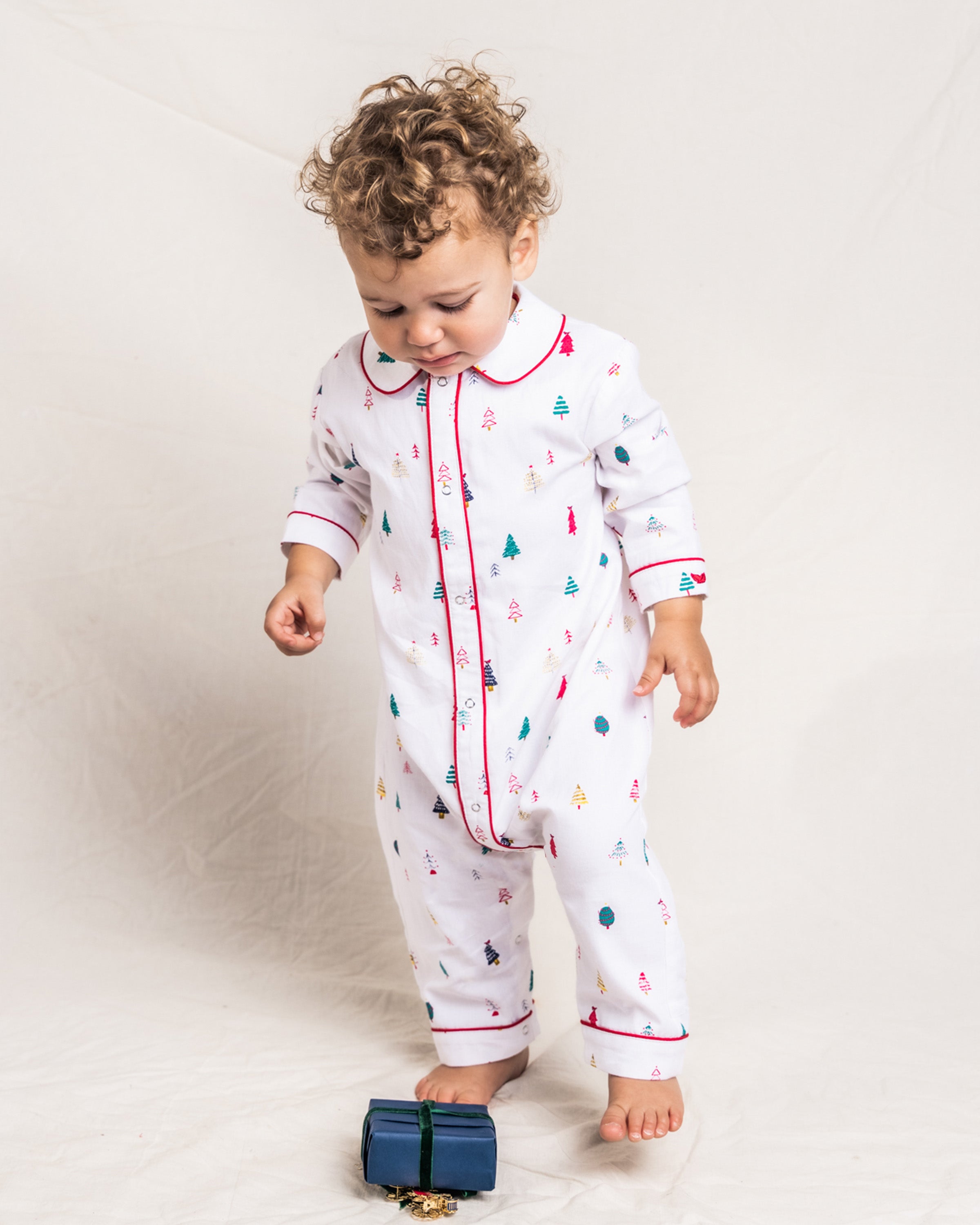 A curly-haired toddler stands in a festive white Babys Twill Cambridge Romper in Merry Trees by Petite Plume, gazing at a small blue gift box. The light, plain background highlights the brushed fabric and cozy, flame-retardant attire with Christmas trees and holiday motifs.
