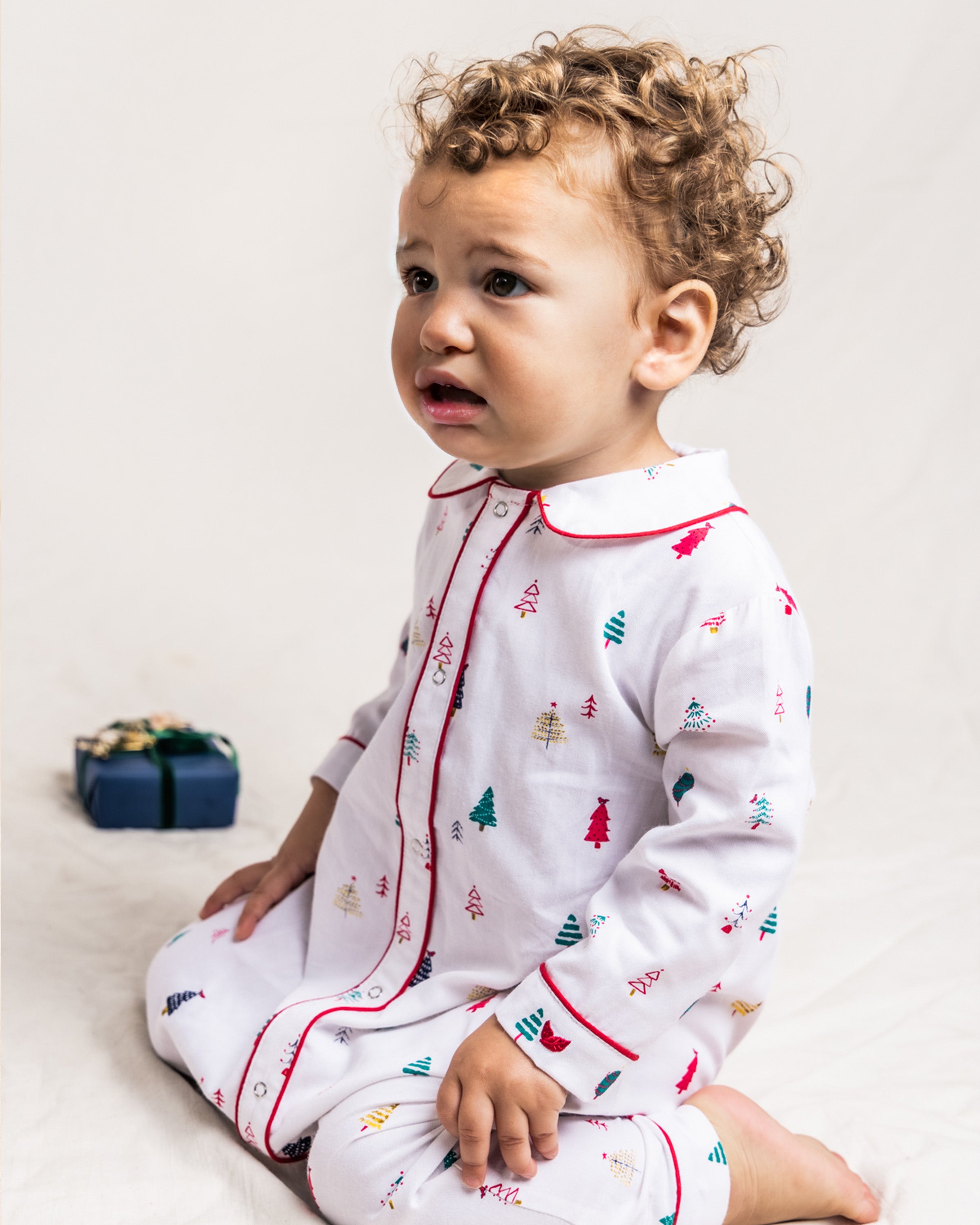 A curious toddler with curly hair sits in cozy Babys Twill Cambridge Romper in Merry Trees by Petite Plume, adorned with small Christmas trees. Next to them lies a small, wrapped gift with a bow on the floor.