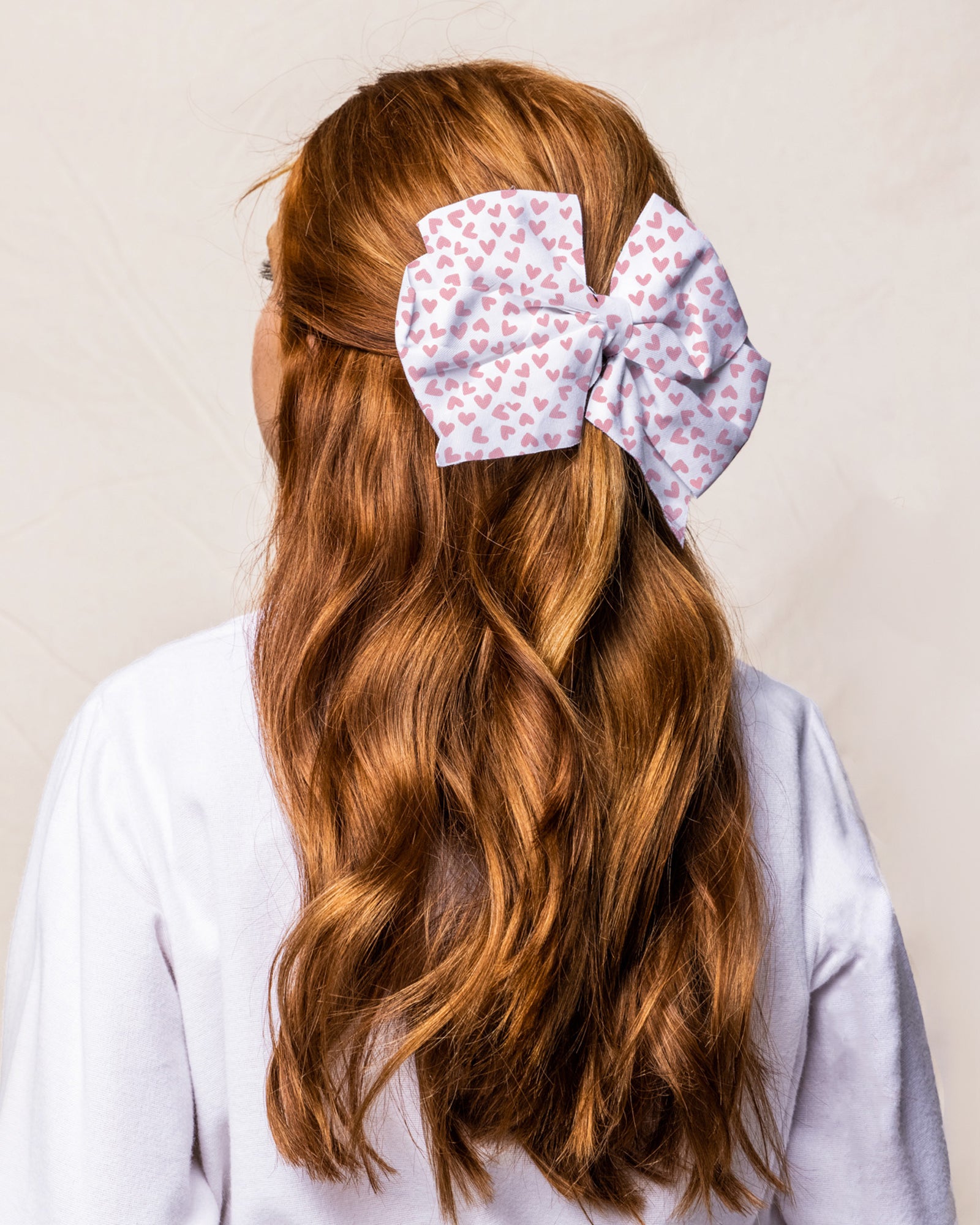 A person with long, wavy red hair, facing away in a white top against a plain background, is wearing the Petite Plume Girls Hair Bows in Sweethearts—a large white bow adorned with a pink heart pattern, ideal for festive occasions.