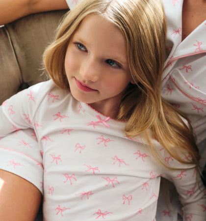 Young girl with long blonde hair wearing white pajamas with pink bow patterns, resting against an adult in matching pajamas, sitting on a beige sofa and looking slightly to the side with a gentle expression.