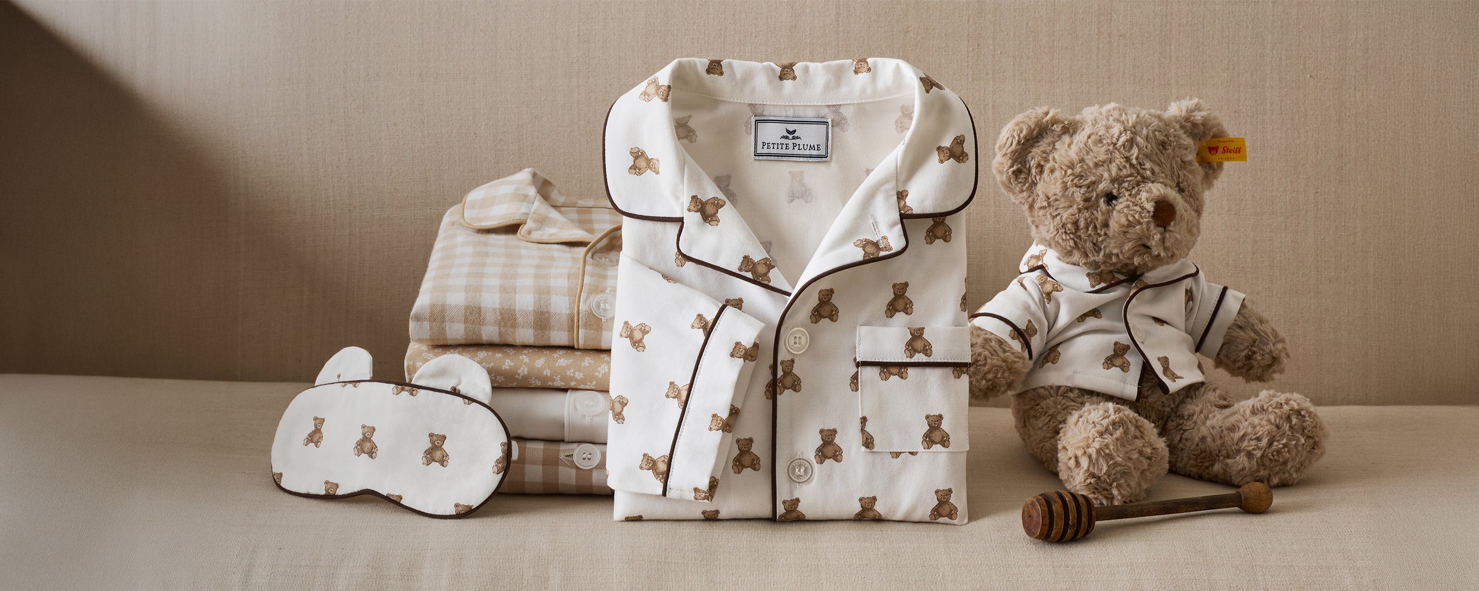 A set of childrens pajamas with a teddy bear print is neatly folded beside matching bedding and an eye mask. A teddy bear wearing similar pajamas sits nearby, next to a honey dipper and a small honey pot.