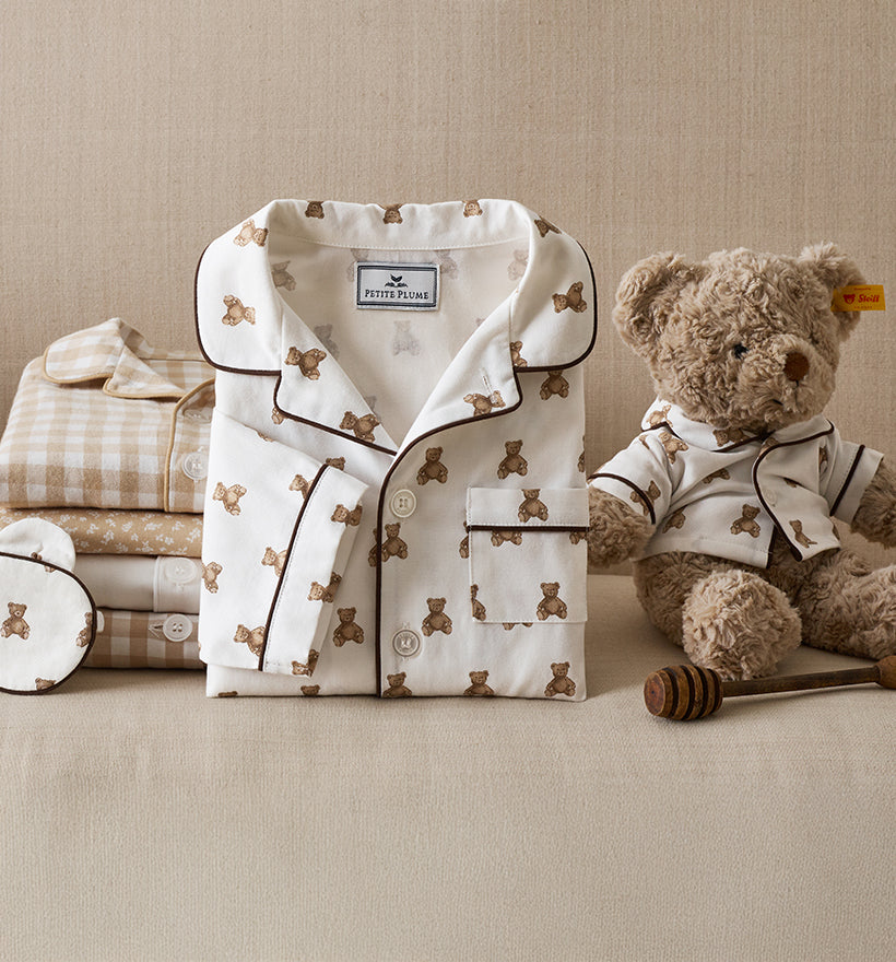 A neatly folded set of children’s pajamas with a teddy bear print is displayed next to matching fabric items and a plush teddy bear wearing similar pajamas, all set against a neutral beige background.