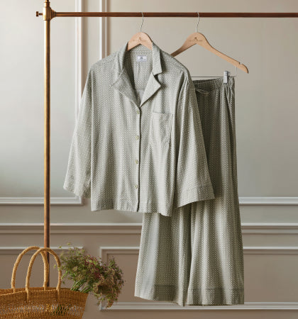 A set of light gray patterned pajamas with a button-up shirt and wide-leg pants hangs on wooden hangers from a gold clothing rack; a woven basket with flowers sits nearby against an elegant white wall.