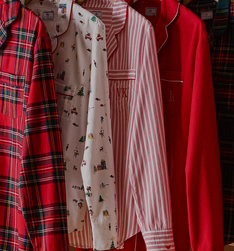 Four long-sleeve pajama tops hang side by side: red plaid, white with holiday prints, red and white stripes, and solid red, each with a front pocket and piping detail on the collar and pocket.