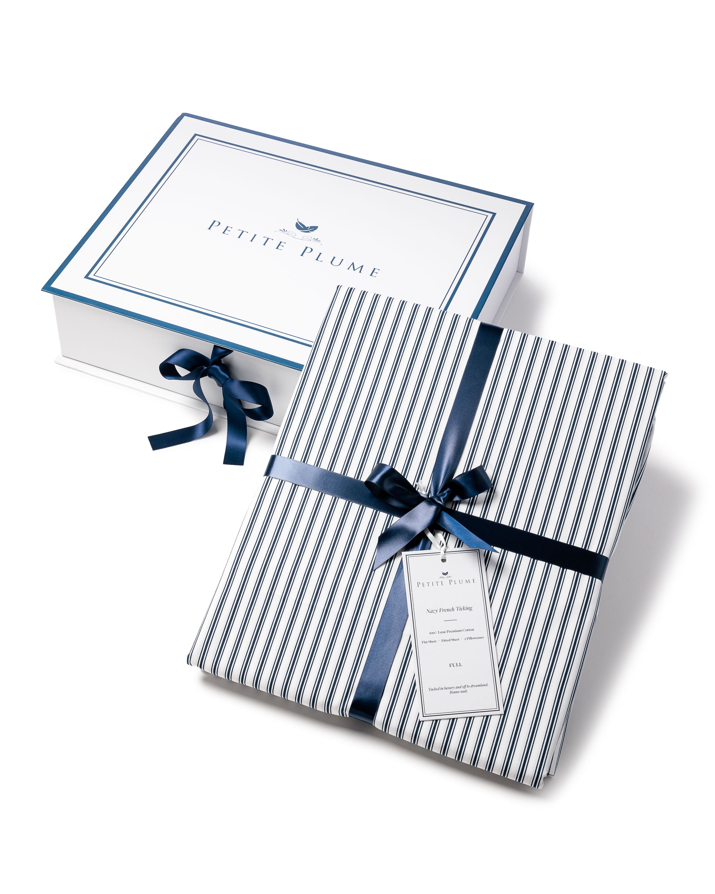 A neatly wrapped package features a navy blue and white striped pattern with a dark blue ribbon and tag, elegantly placed in front of a white box bordered in blue with PETITE PLUME at the center, echoing the luxe feel of Petite Plumes Luxe Premium Cotton Bed Sheets in Navy French Ticking.