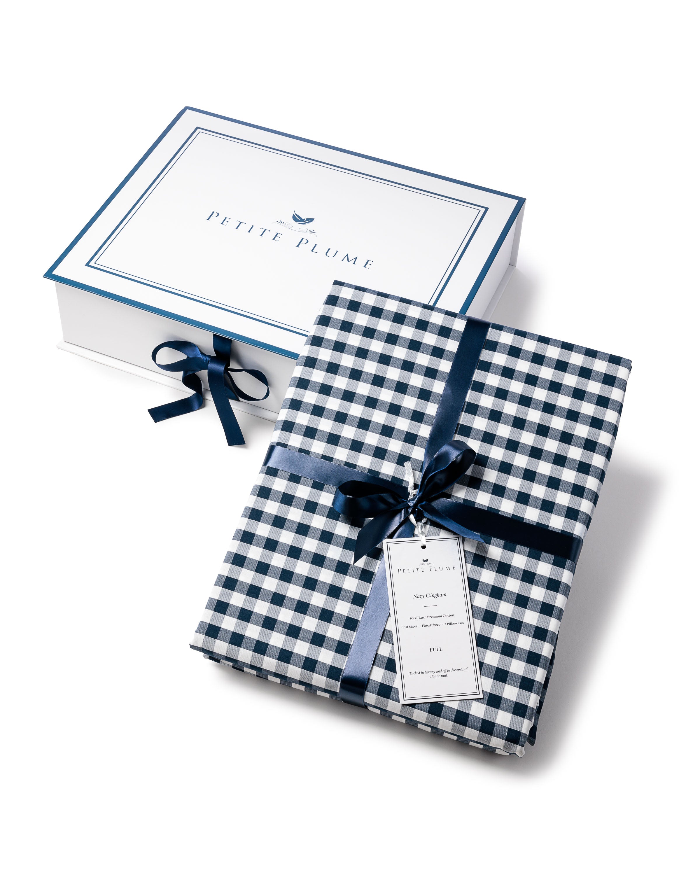 Luxe Premium Cotton Navy Gingham Bed Sheets neatly folded and tied with a dark blue ribbon exude elegance. Placed beside a white box featuring a blue border and the Petite Plume logo, the tag provides product details in style.