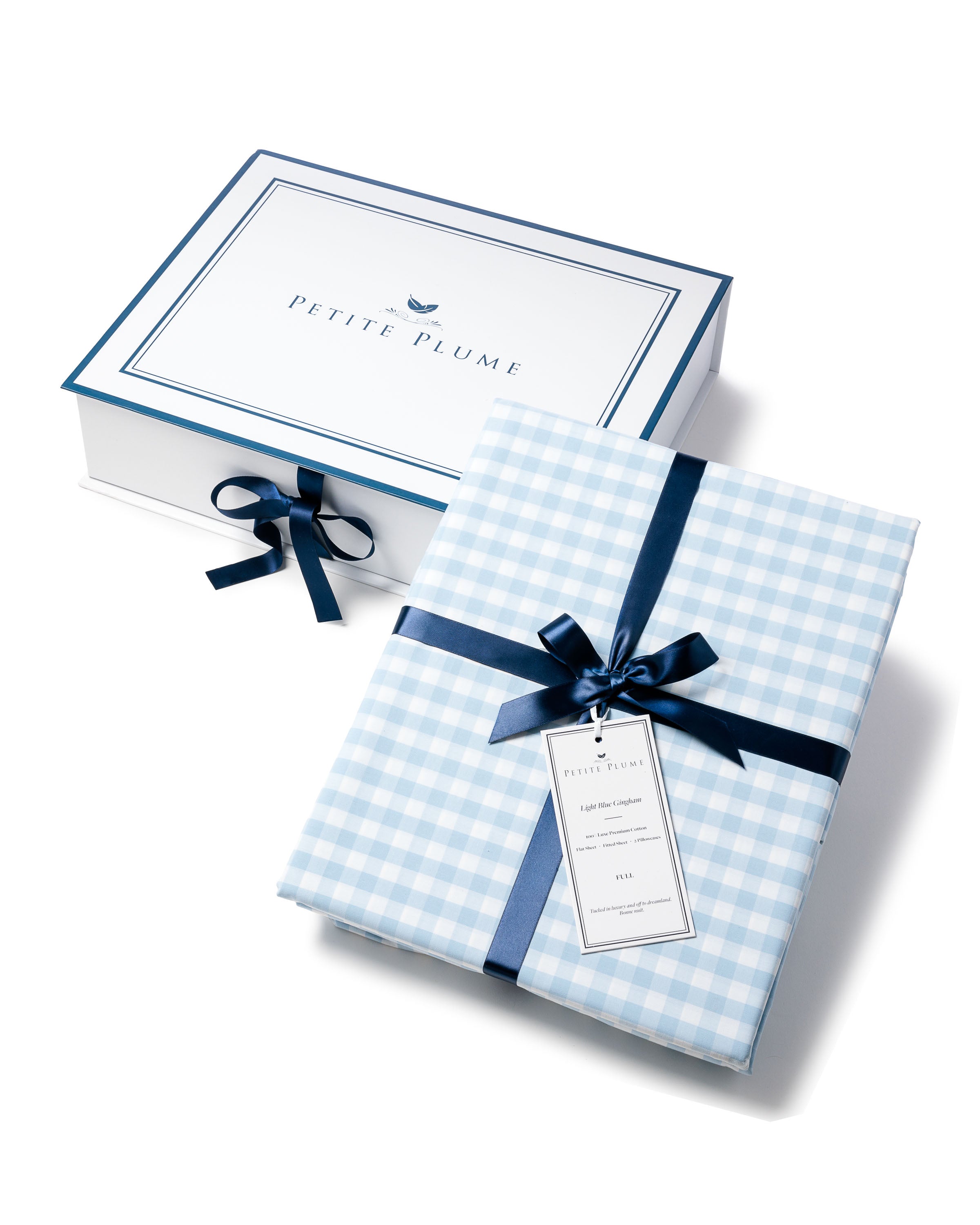 A folded Luxe Premium Cotton Light Blue Gingham Bed Sheet, tied with a dark blue ribbon and label, rests elegantly before a Petite Plume box adorned with a matching ribbon, all against a pristine white background.