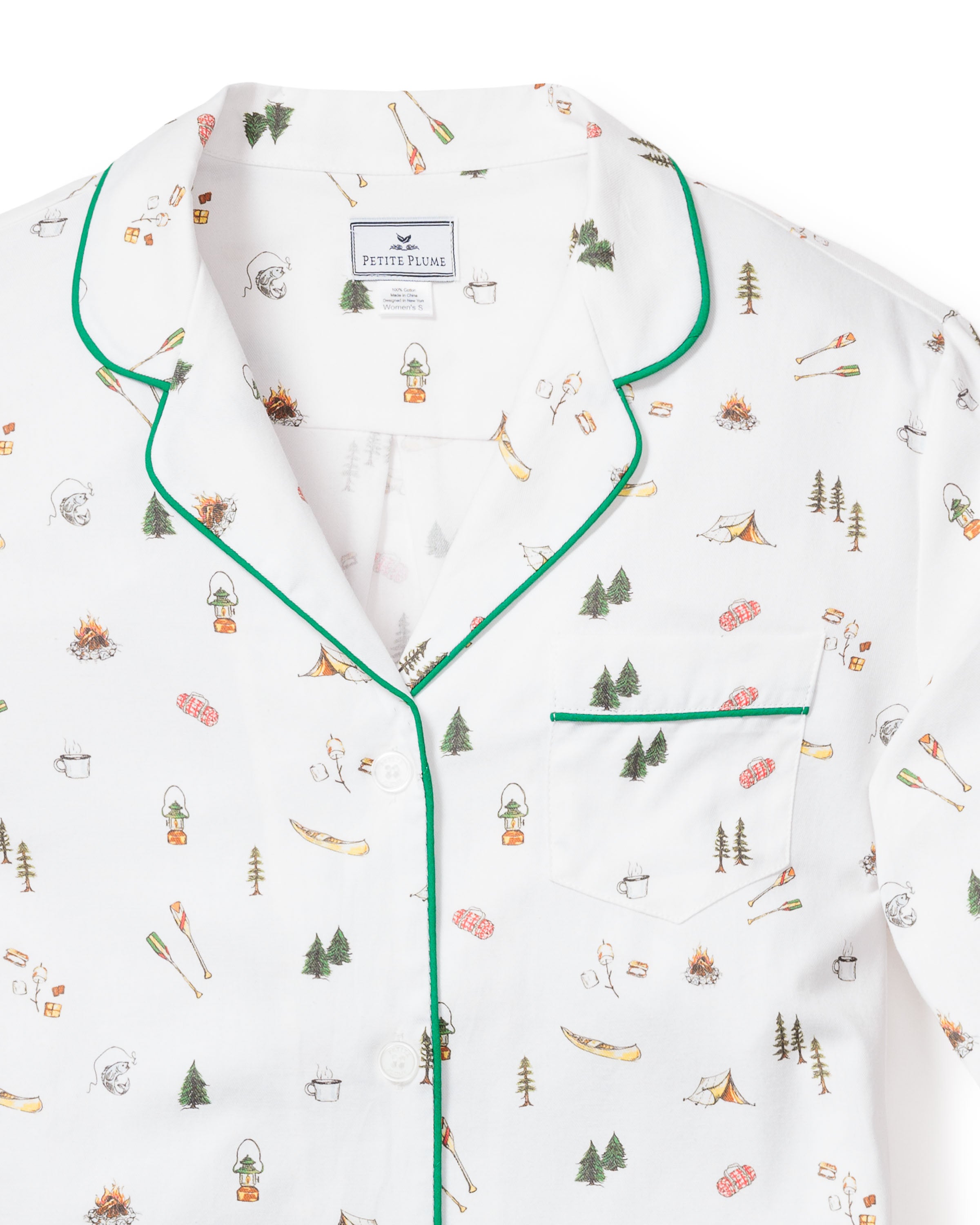 The Womens Twill Pajama Set in The Great Outdoors by Petite Plume features white cotton with green piping and a playful camping-themed pattern of tents, trees, campfires, and canoes. It includes a buttoned-down top with a chest pocket and the Petite Plume label at the neckline.