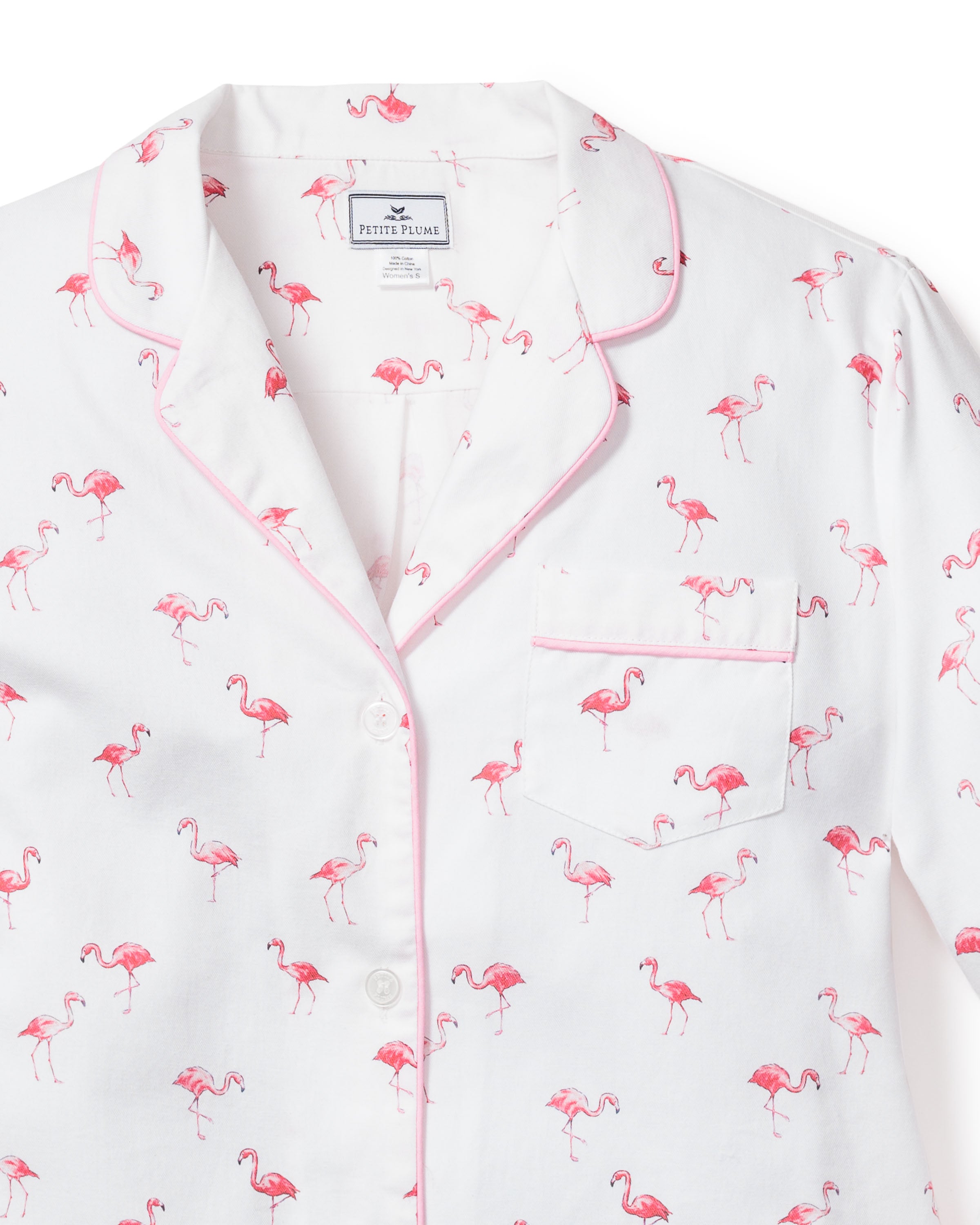 The Womens Twill Pajama Set in Flamingos by Petite Plume features a white top with pink piping and pocket, adorned with small pink flamingos for a tropical touch. It has a notched collar and a visible neckline label, offering luxurious sleepwear vibes.