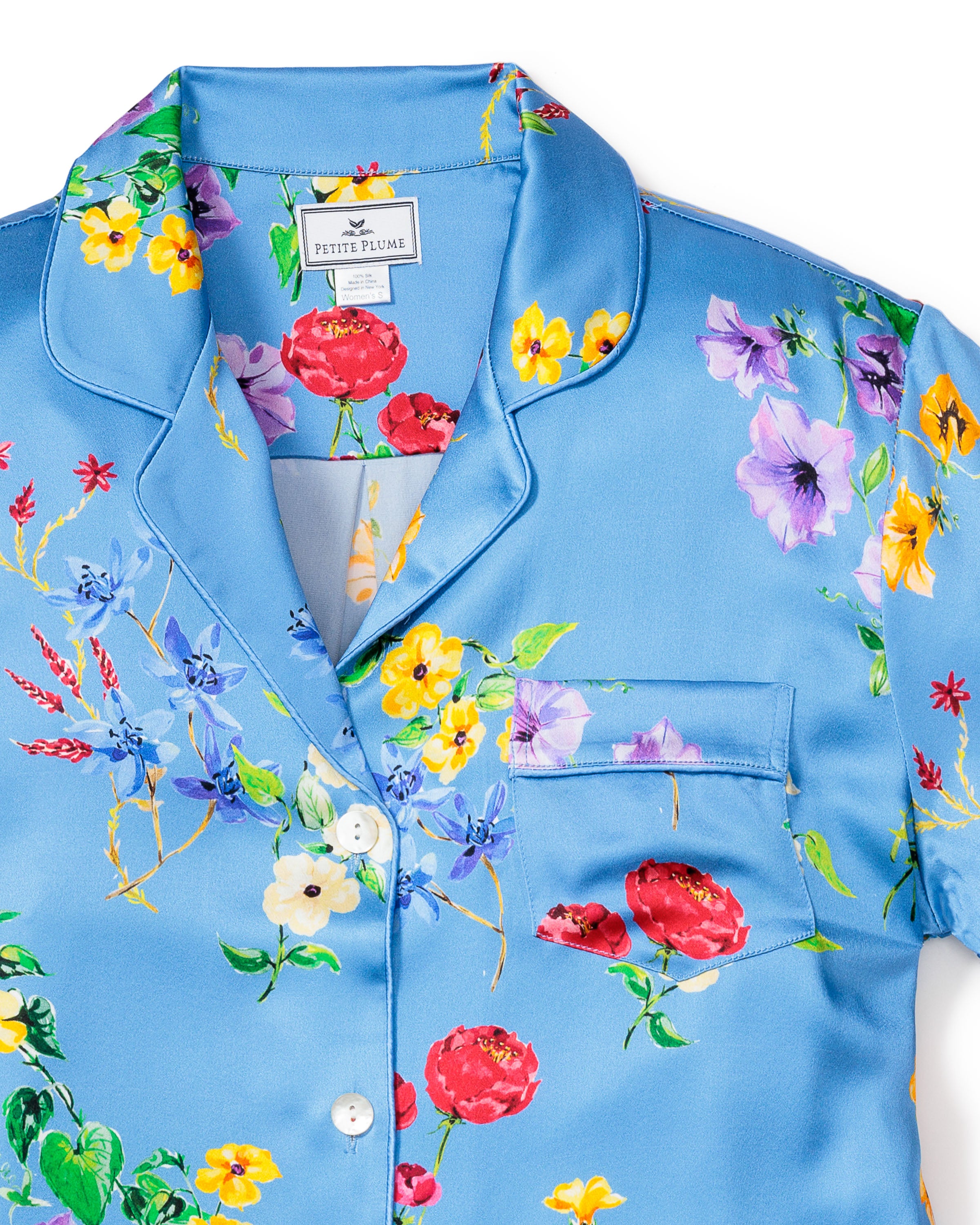 The Womens Silk Pajama Short Set in Brilliant Botanical Azure by Petite Plume is a luxurious top with a vibrant floral pattern of red, yellow, purple, and blue flowers. It features a collar, chest pocket, white buttons, and has a visible neckline label.