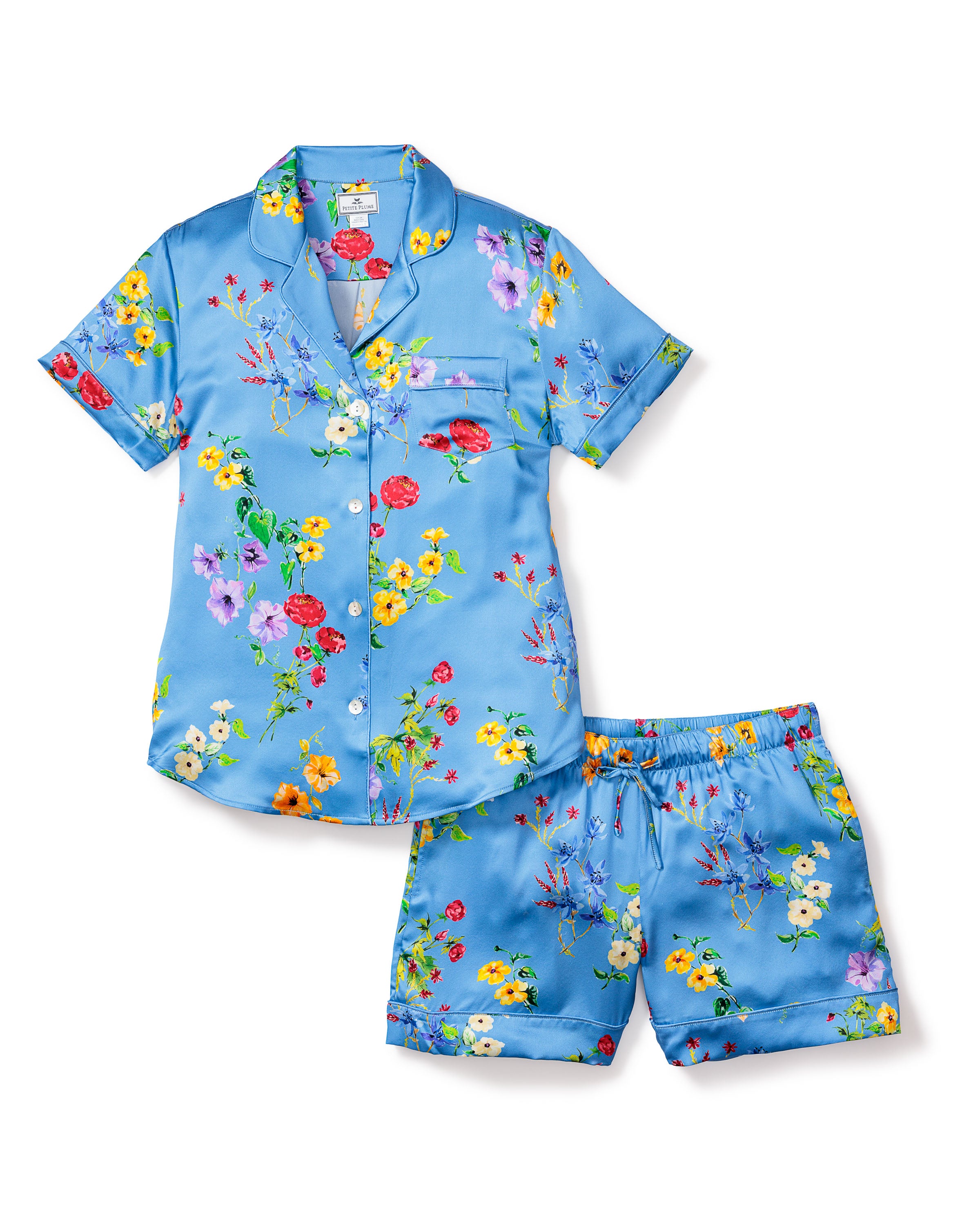 The Womens Silk Pajama Short Set in Brilliant Botanical Azure by Petite Plume features luxurious Mulberry silk with vibrant red, yellow, purple, and white floral prints. It has short sleeves, shorts, a button-down front, and a charming pocket on the left side.