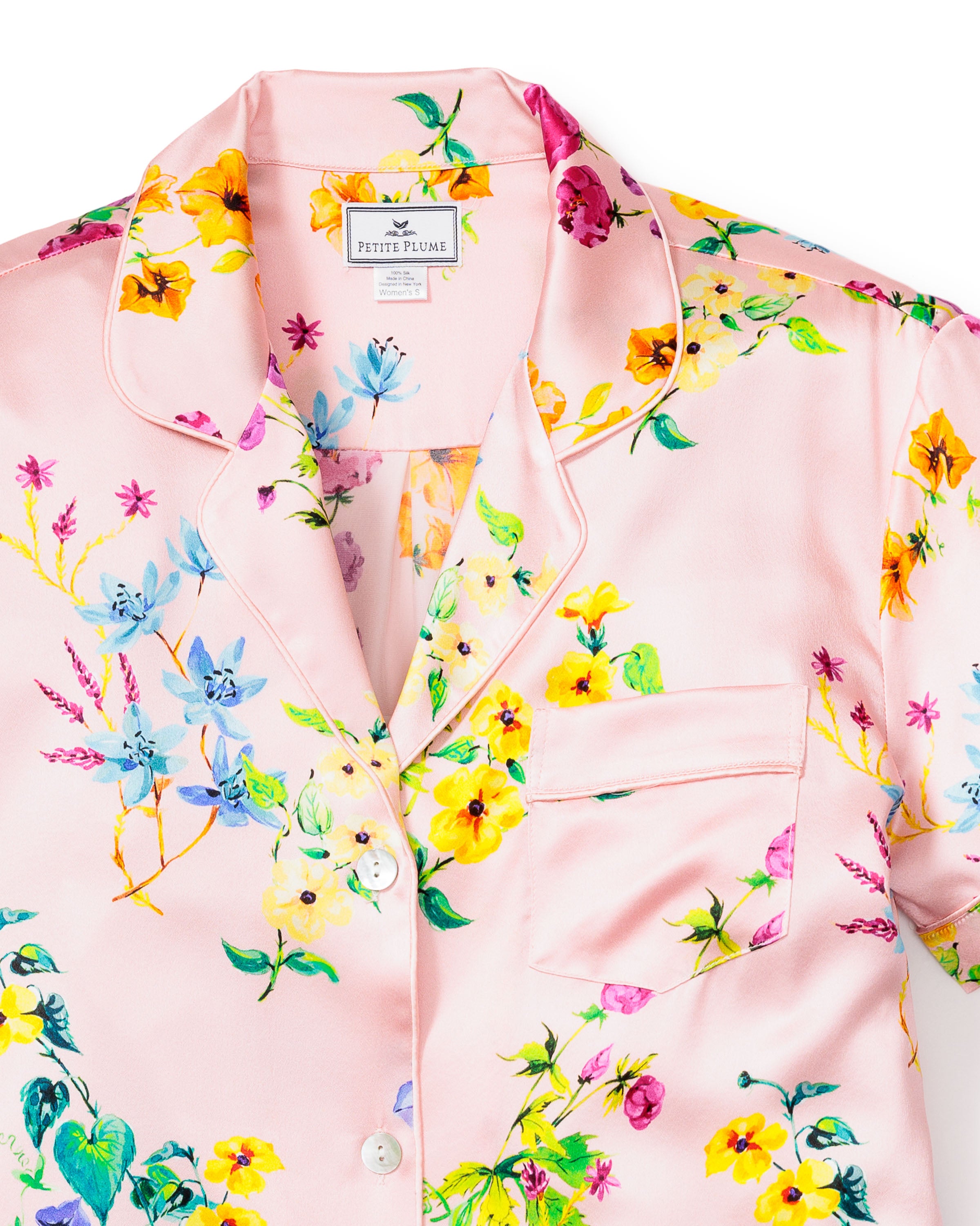 A Petite Plume Womens Silk Pajama Top in Blush Brilliant Botanical, featuring a notched collar, chest pocket, and front buttons. The vibrant print displays yellow, blue, and magenta flowers with green leaves. A label graces the neckline.