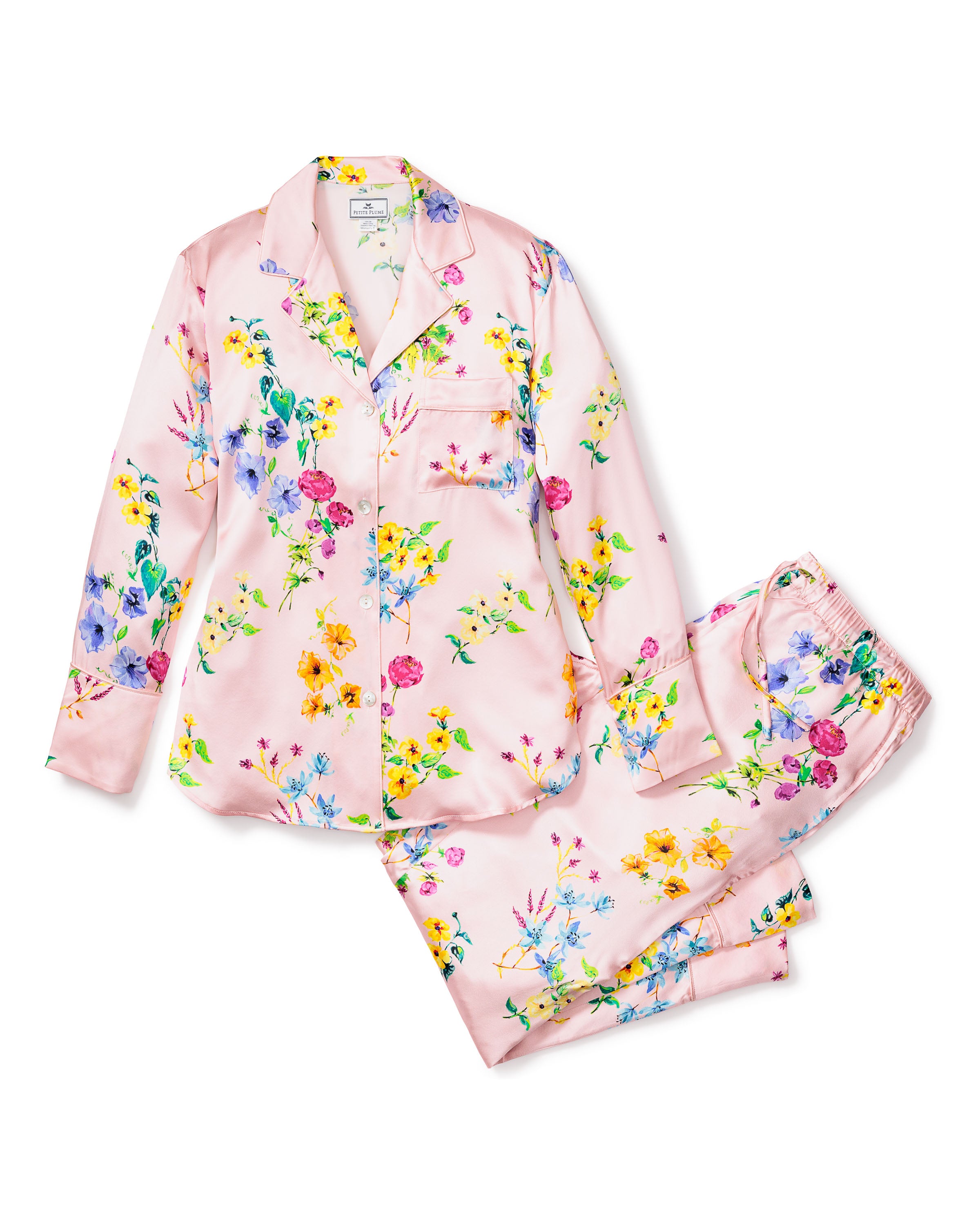 The Womens Silk Pajama Set in Blush Brilliant Botanical by Petite Plume features luxury sleepwear with a long-sleeve button-up shirt with a chest pocket and matching pants with an elastic waistband, adorned in vibrant yellow, purple, and pink floral patterns.