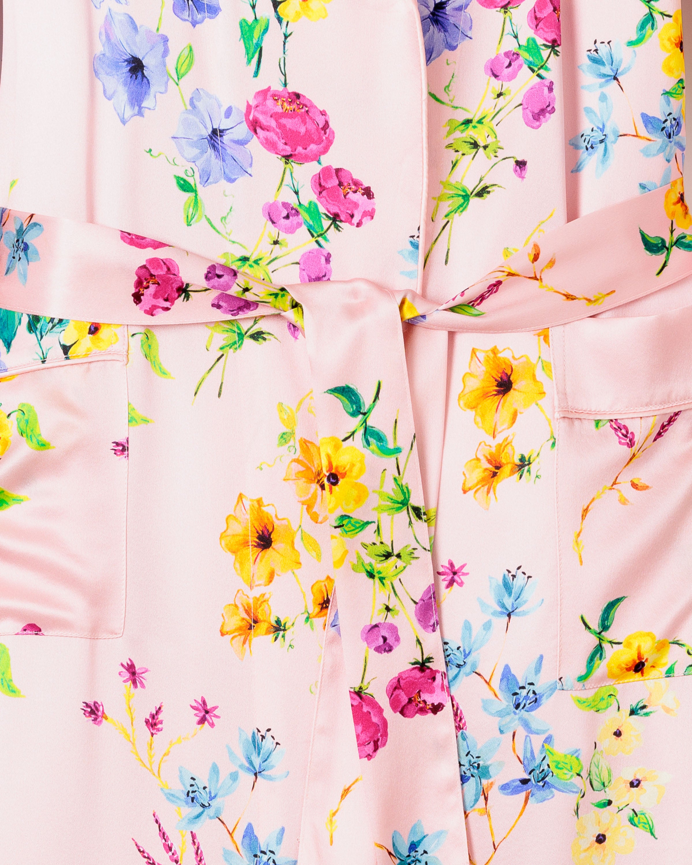 A close-up of Petite Plumes Womens Silk Robe in Blush Brilliant Botanical, displaying a floral pattern with yellow, blue, and pink hues. The garment features pockets and a tied belt on silk canvas fabric adorned with blossoms and leaves.