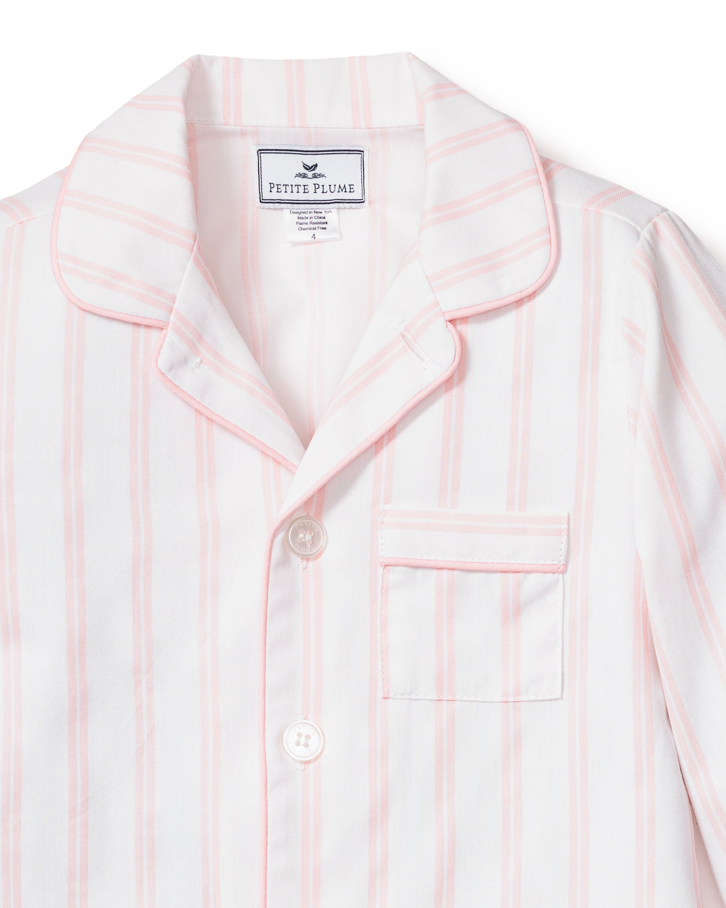 Close-up of a luxurious Kids Twill Pajama top in pink and white stripes. It has a notched collar, chest pocket, and two buttons. The label reads Petite Plume. The cotton fabric looks soft and smooth.
