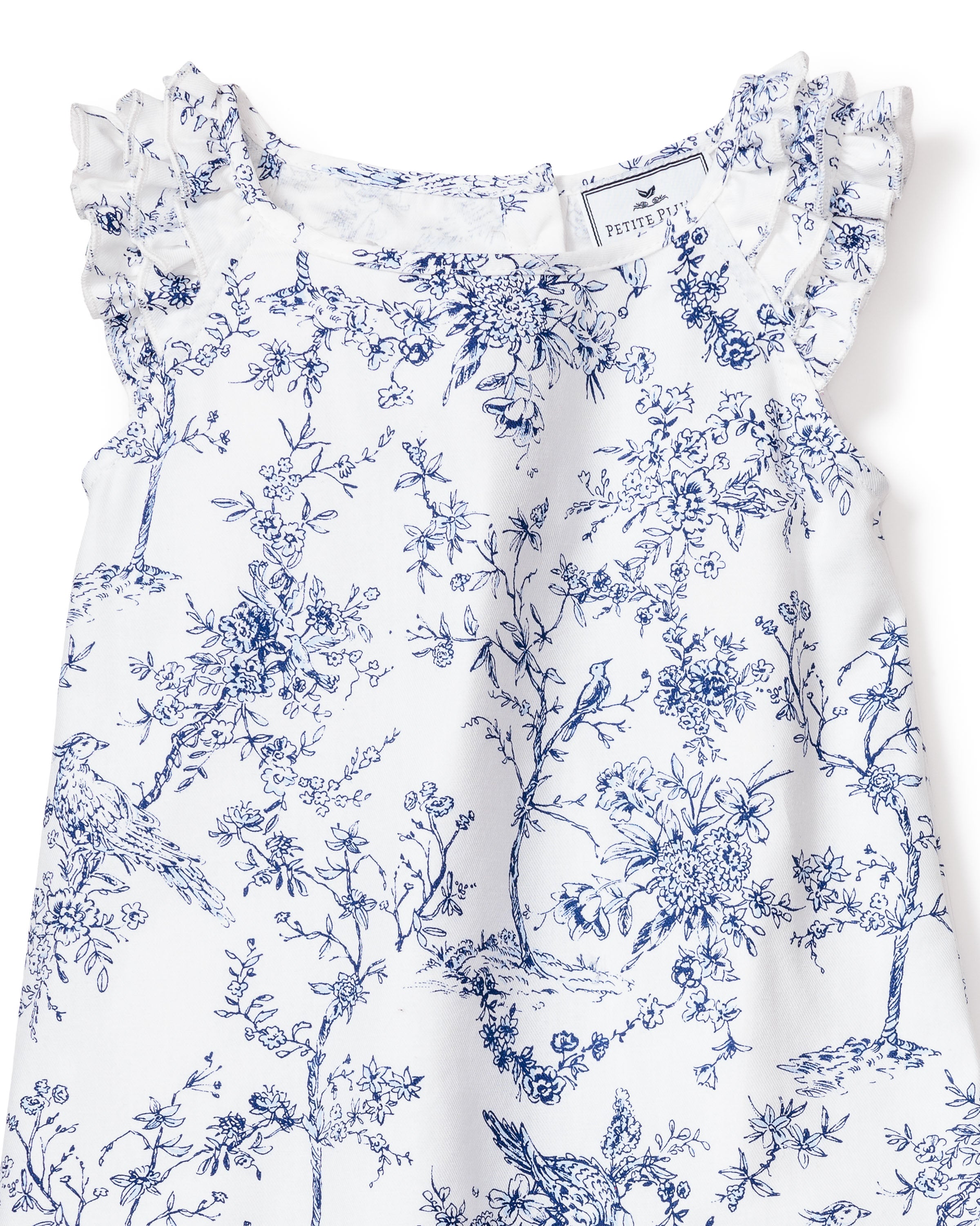 The Girls Twill Amelie Short Set in Timeless Toile by Petite Plume boasts a sleeveless white dress with blue floral and bird prints. Made from luxury cotton, it features ruffled shoulders, a round neckline, and a small tag near the neck that adds charm.