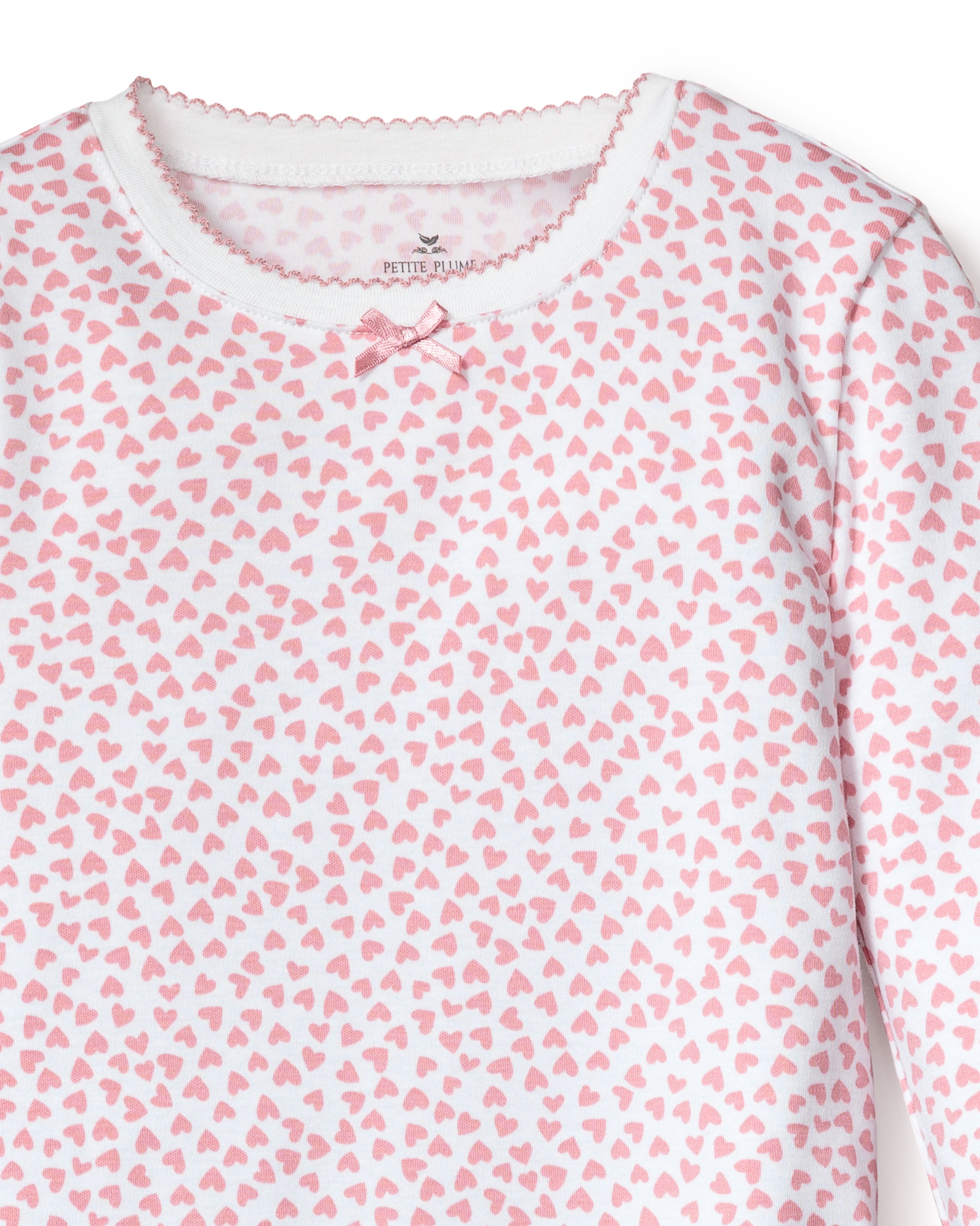The Kids Pima Snug Fit Pajama Set in Sweethearts by Petite Plume features a luxurious white long-sleeve top adorned with a charming pink heart pattern. Crafted from soft Pima cotton, it includes a small pink bow and scalloped trim at the neckline for an effortlessly stylish look.
