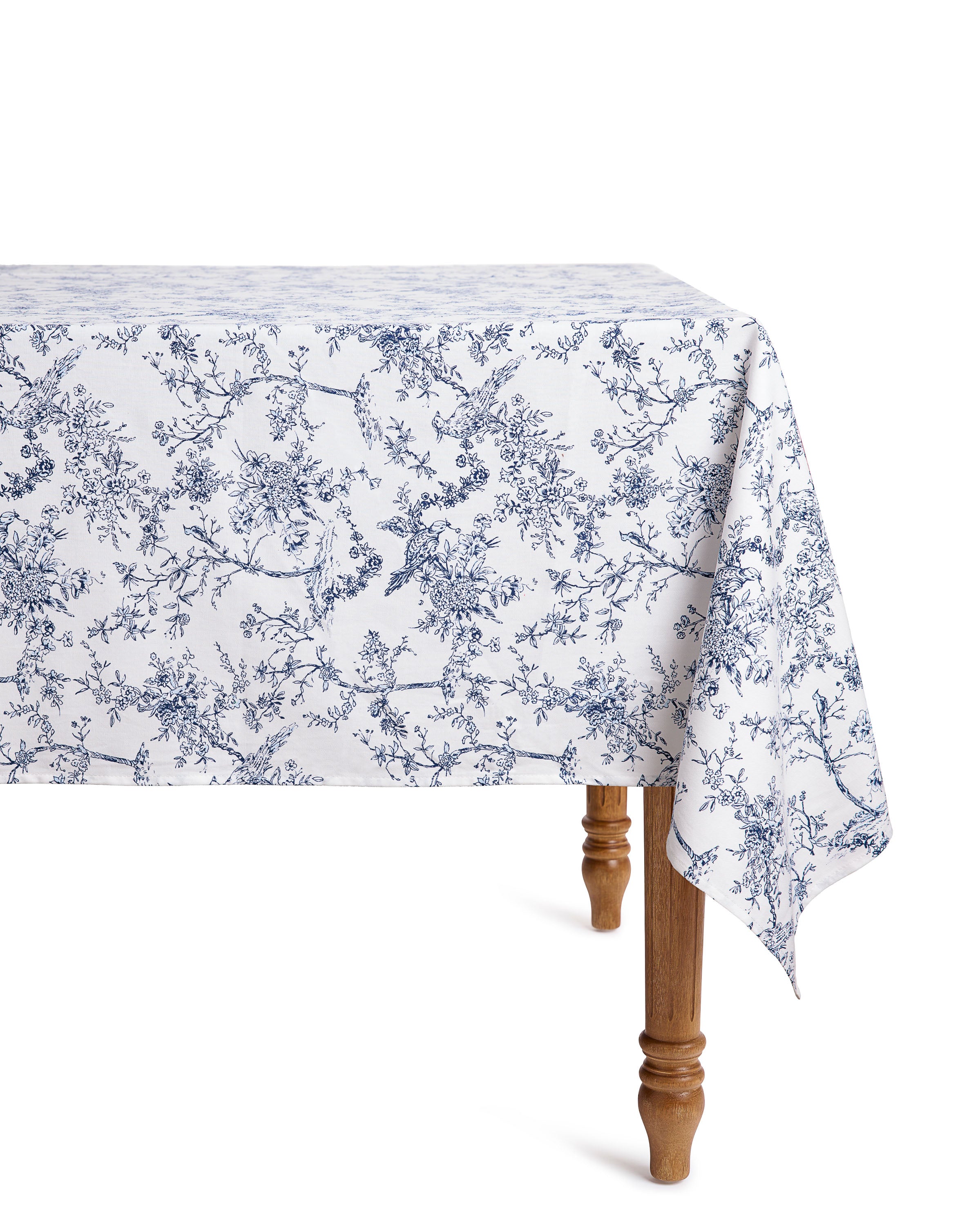 A wooden table with cotton linens features a Signature Twill Tablecloth from Petite Plume, showcasing the Timeless Toile print with blue floral patterns. The cloth elegantly drapes over the sides, revealing intricate branches and flowers, while only one leg of the table is visible.