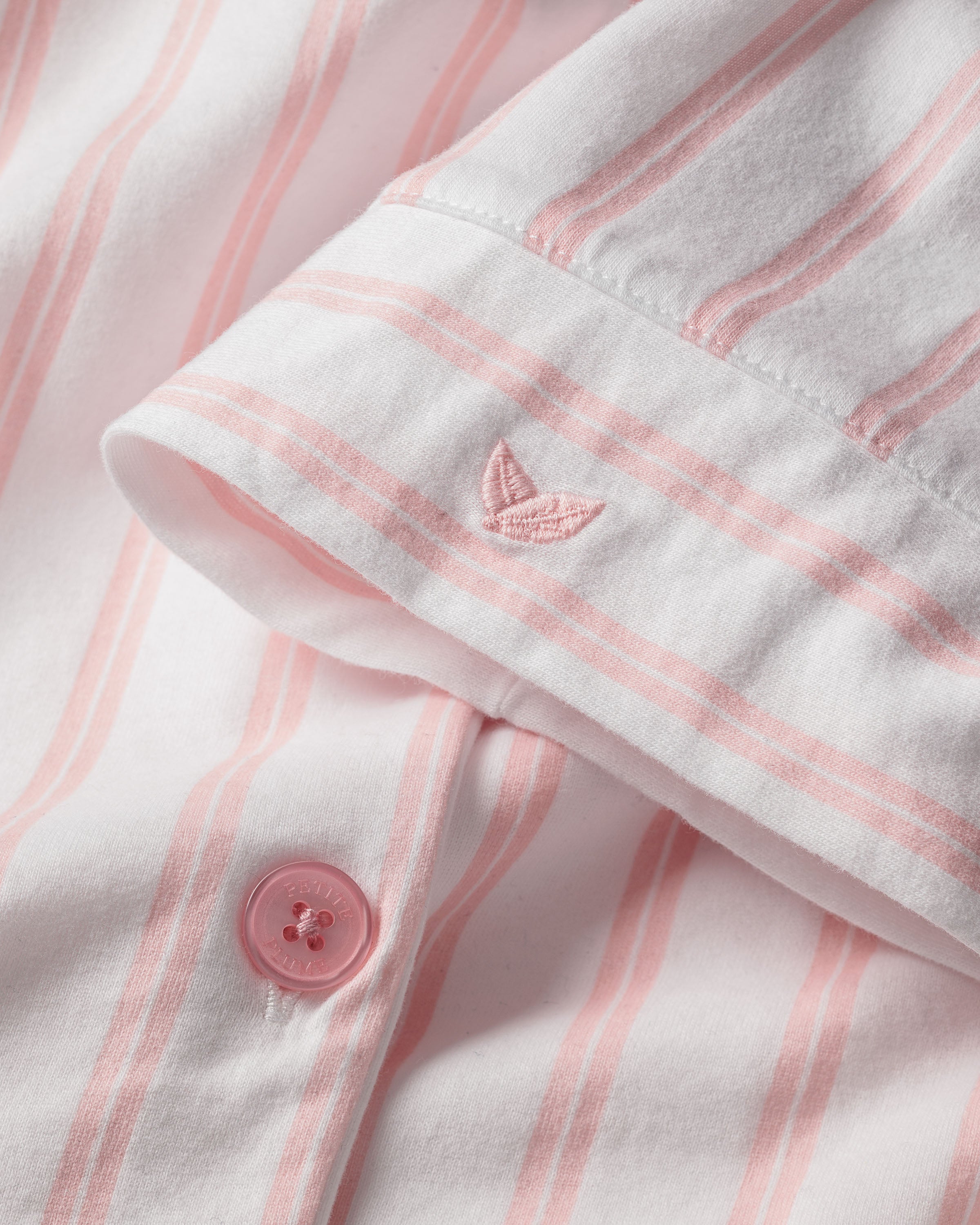 Close-up of the Olivia Wide Leg Pima Pajama Set in Pink Stripe by Petite Plume, featuring a soft white fabric with vertical pink stripes. A pink embroidered plant logo and a pink cuff button enhance these luxurious Peruvian Pima cotton pajamas, perfect for your bedtime ritual.