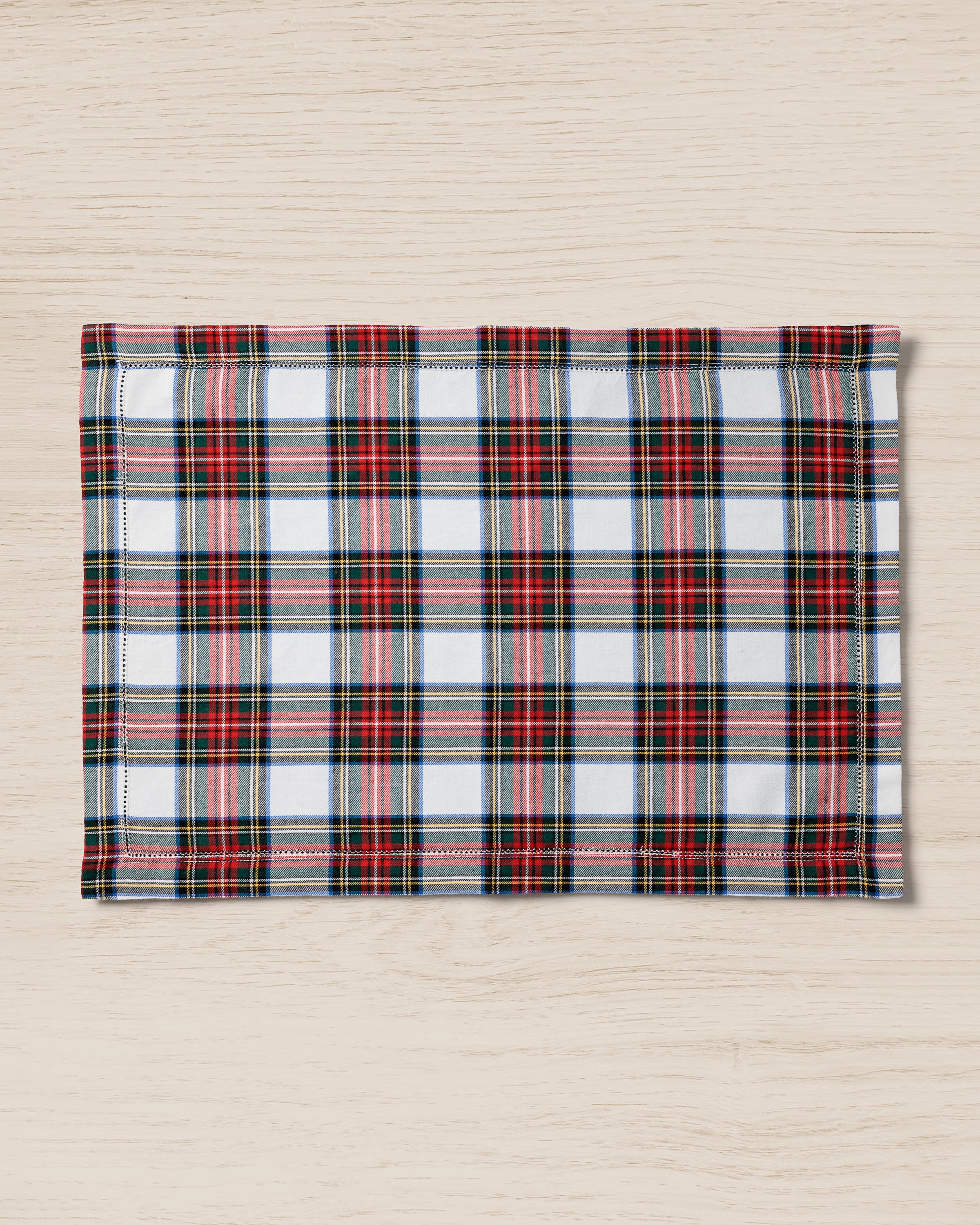 The Signature Twill Placemats in Balmoral Tartan by Petite Plume feature a classic tartan design with red, black, yellow, and green lines on a white background, enhancing your table with versatile prints on a light wooden surface.