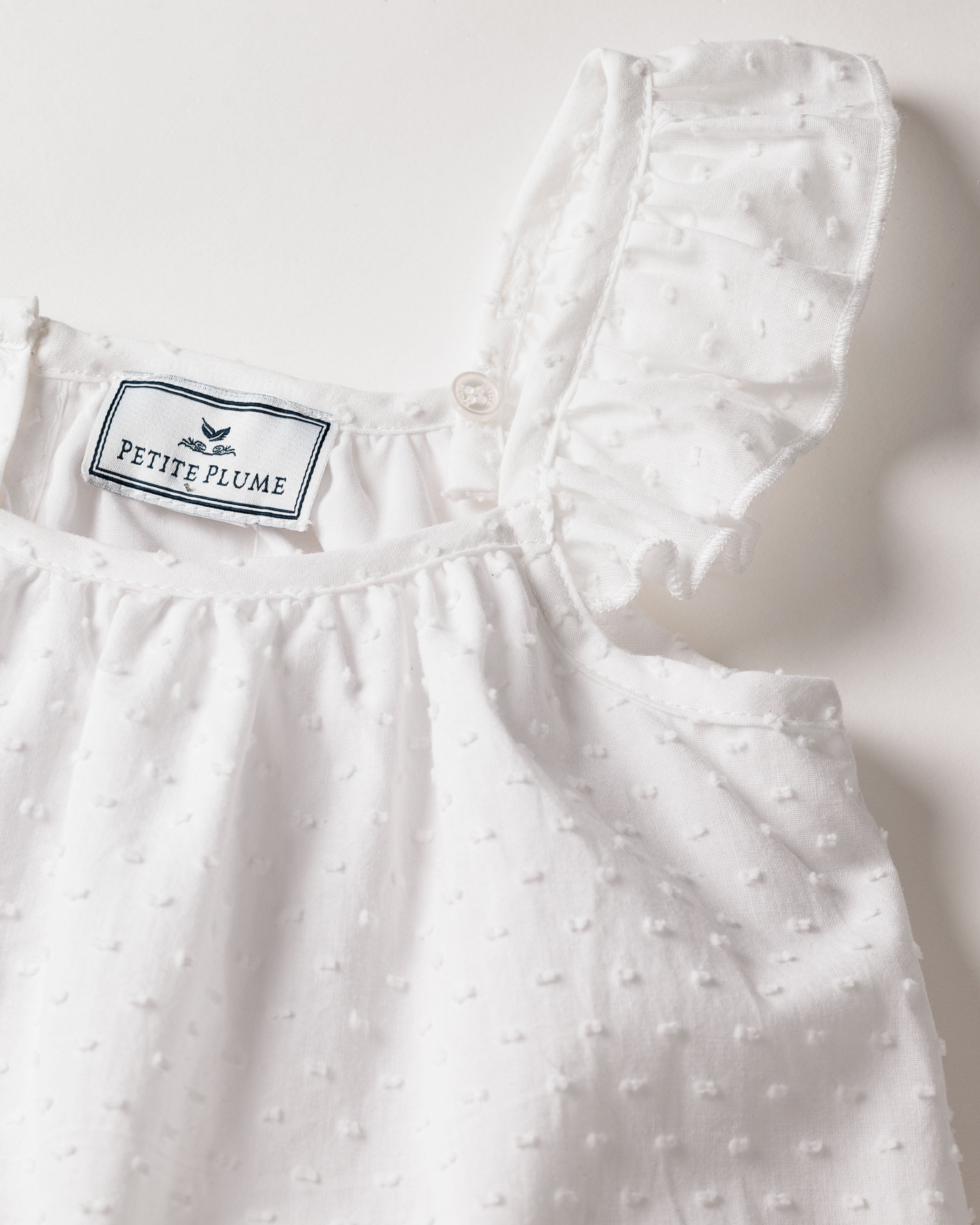 The Girls Swiss Dots Celeste Day Dress in White by Petite Plume features ruffled straps and a textured Swiss Dot Cotton design. This elegant, lightweight garment exudes luxury sleepwear appeal against a plain background.