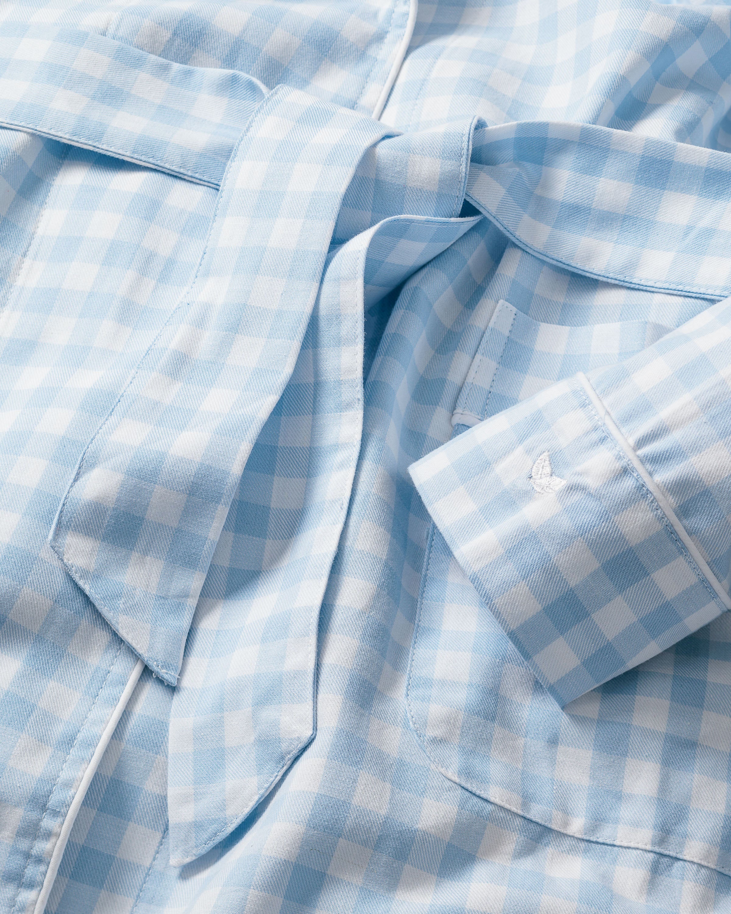 A close-up of Petite Plumes Kids Twill Robe in Light Blue Gingham showcases a folded sleeve and pocket. The chemical-free fabric offers a soft, smooth texture with an evenly distributed checkered pattern for a casual yet classic look.
