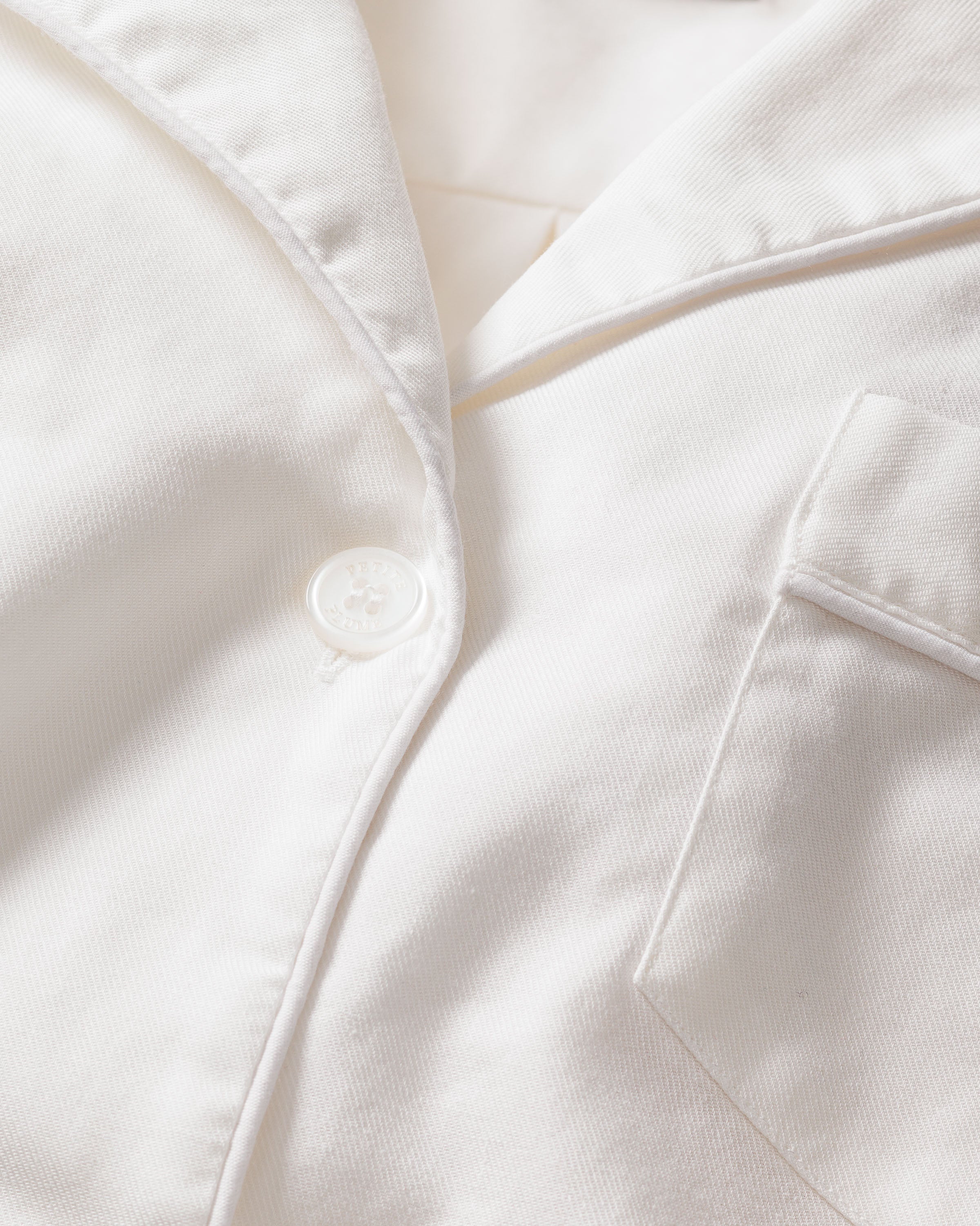 Close-up of the Womens Twill Pajama Short Set in White by Petite Plume, showcasing a button, lapel, and pocket. The smooth texture highlights exquisite stitching and craftsmanship with finest quality cotton.