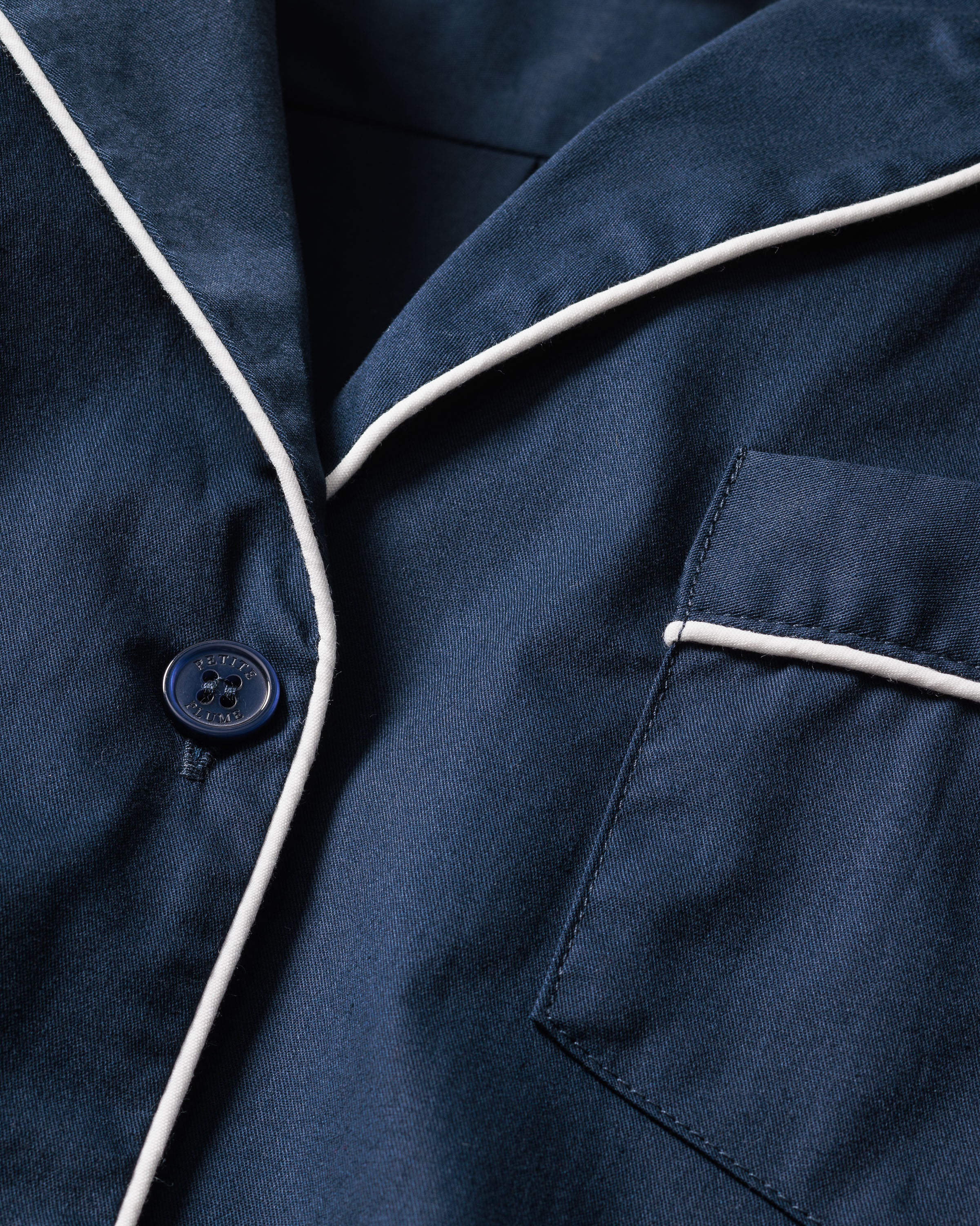 A close-up of navy fabric with a button and white piping outlines the Womens Navy Twill Long Sleeve Short Set from Petite Plume. The cotton twill material includes a right-side pocket, implying its part of a cozy pajama set with possible moisture-wicking properties for comfort.