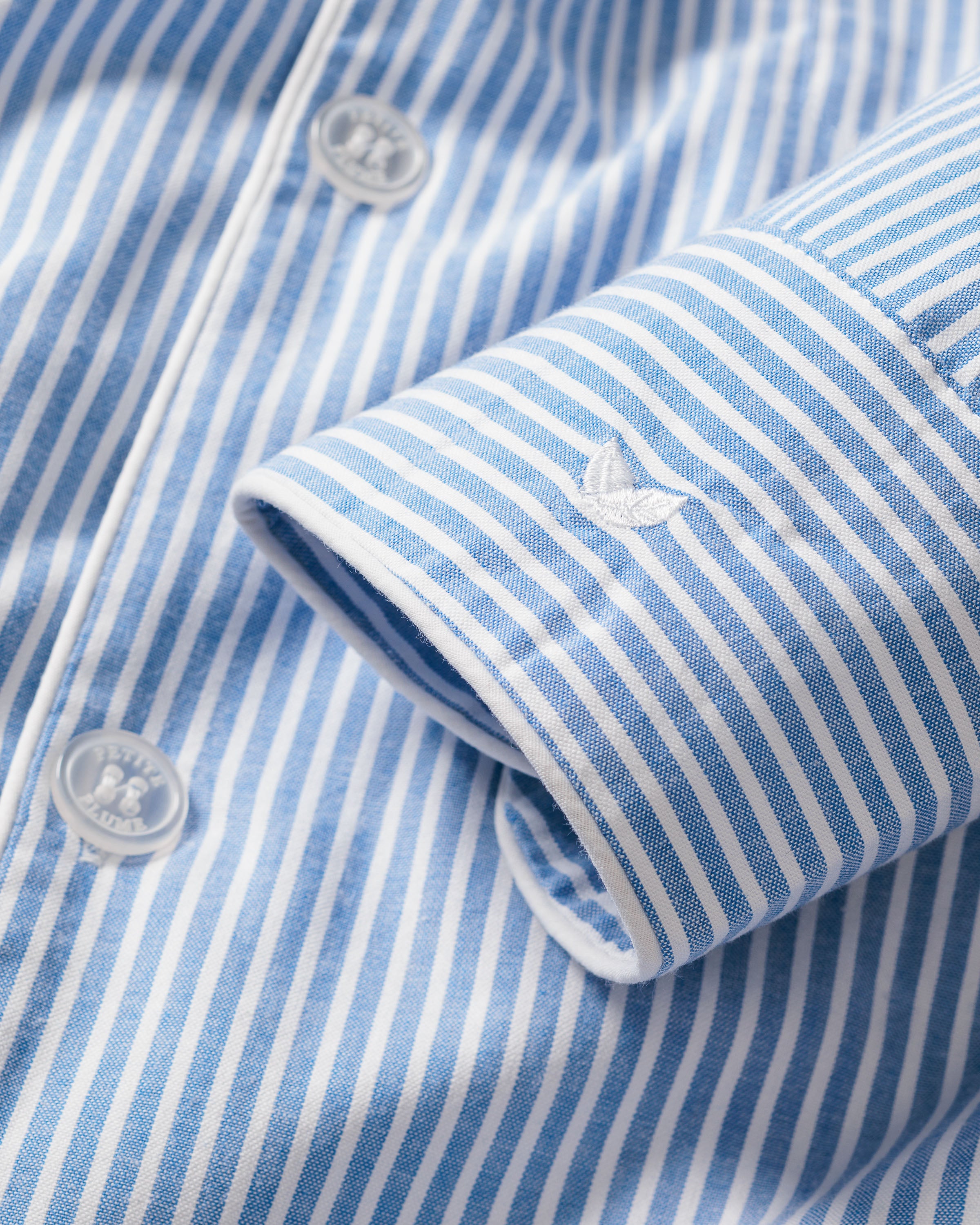 The Mens Twill Pajama Set by Petite Plume features a close-up of a French Blue striped seersucker shirt, showcasing visible buttons, cuff stitching, and an embroidered motif. The crisp 100% cotton fabric gives it a classic pajama set look.