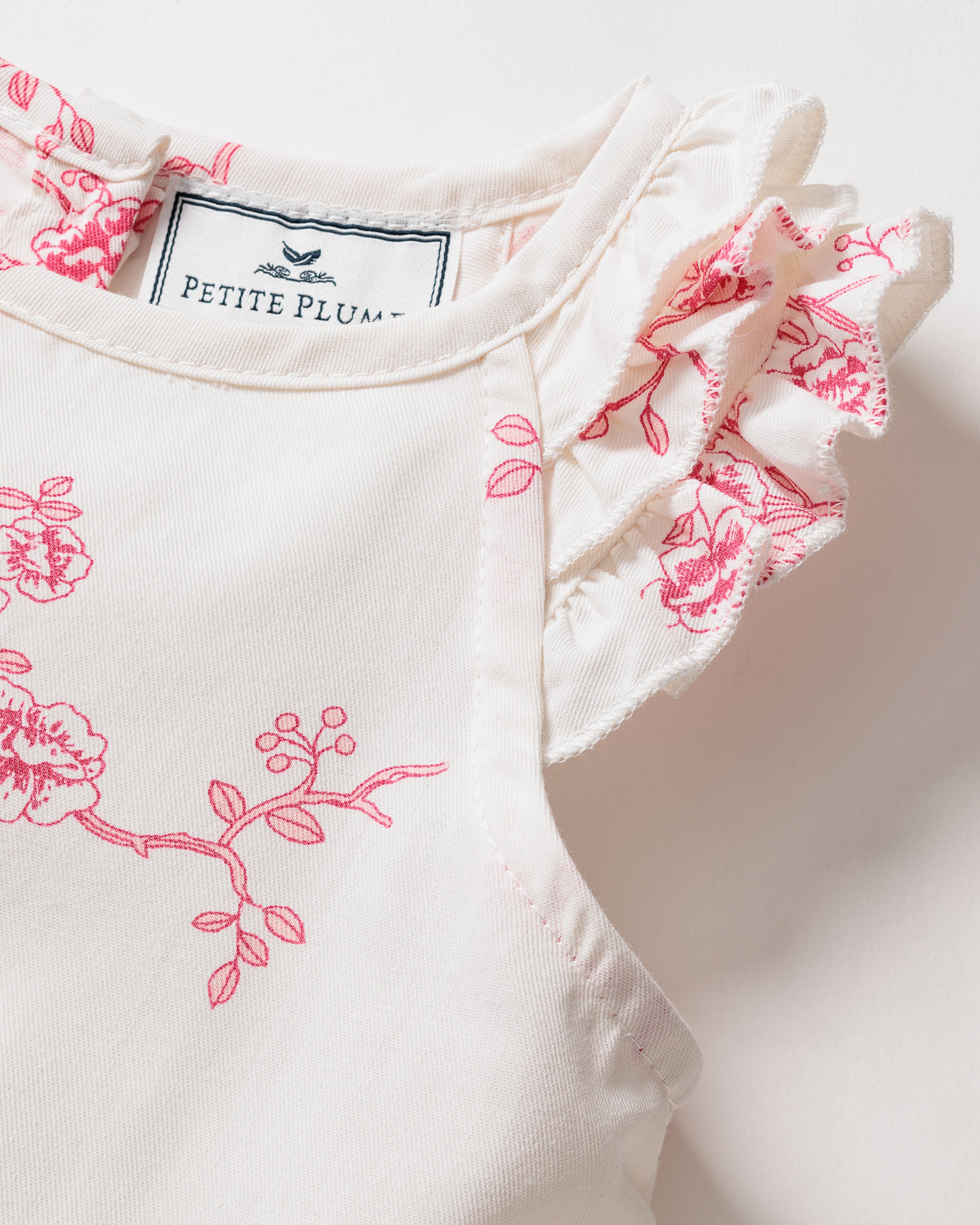 Close-up of the Girls Twill Amelie Nightgown in English Rose Floral shows cream fabric with vintage-style pink florals and ruffled sleeves. A small Petite Plume label with a bird illustration highlights the elegance of luxurious sleepwear.