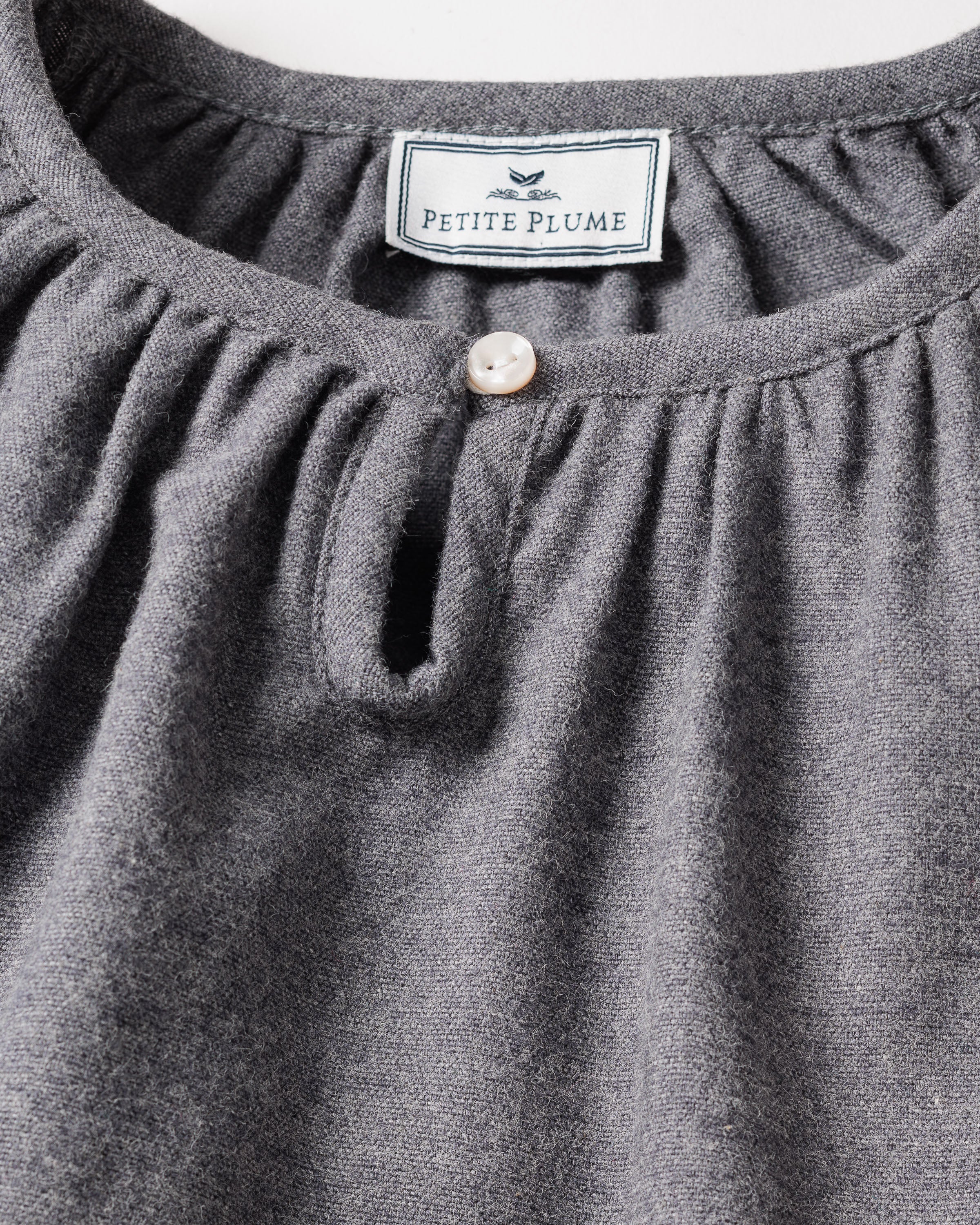 Close-up of a grey nightgown showcasing a round neckline and pearl-like button. Inside, the label reads Petite Plume with a bird graphic. Made from brushed cotton flannel, its texture highlights the softness and chemical-free comfort of the Girls Flannel Delphine Nightgown in Grey by Petite Plume.