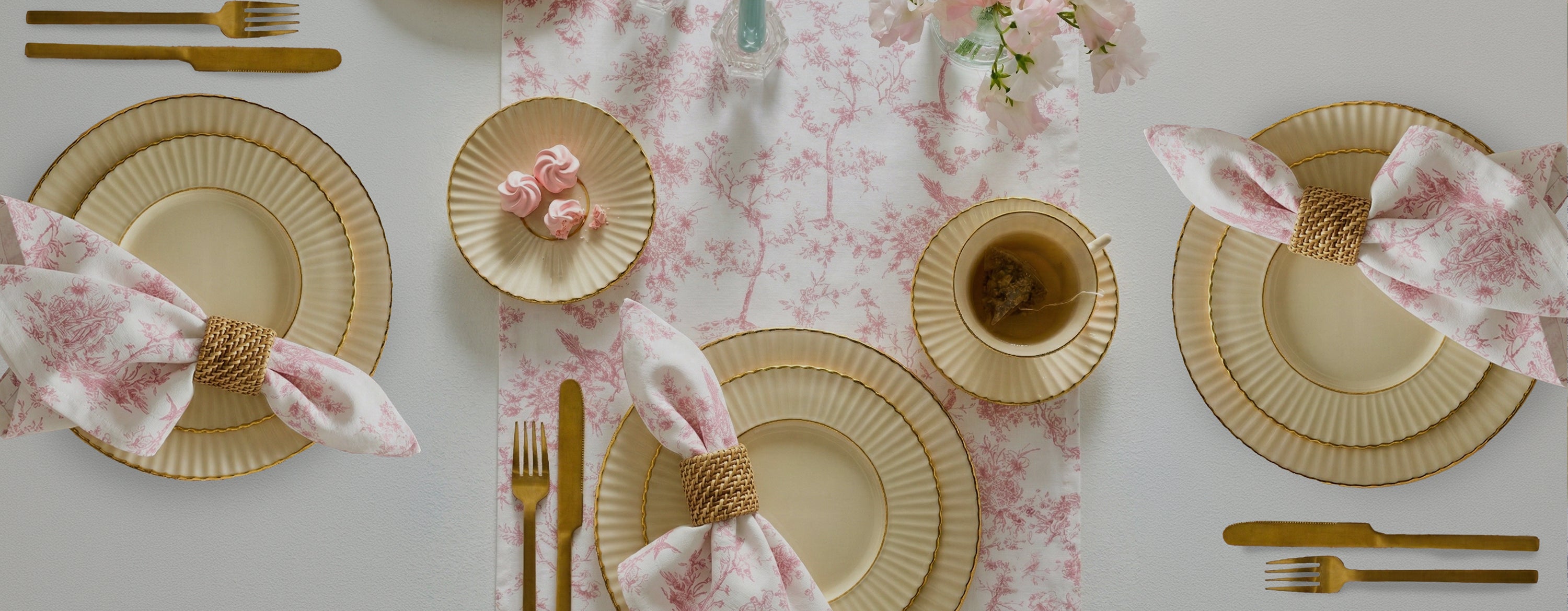 An elegant table setting with cream and gold plates, gold cutlery, teacups, and pink-patterned napkins in woven rings on a white and pink floral table runner. Small pastries sit on side plates.