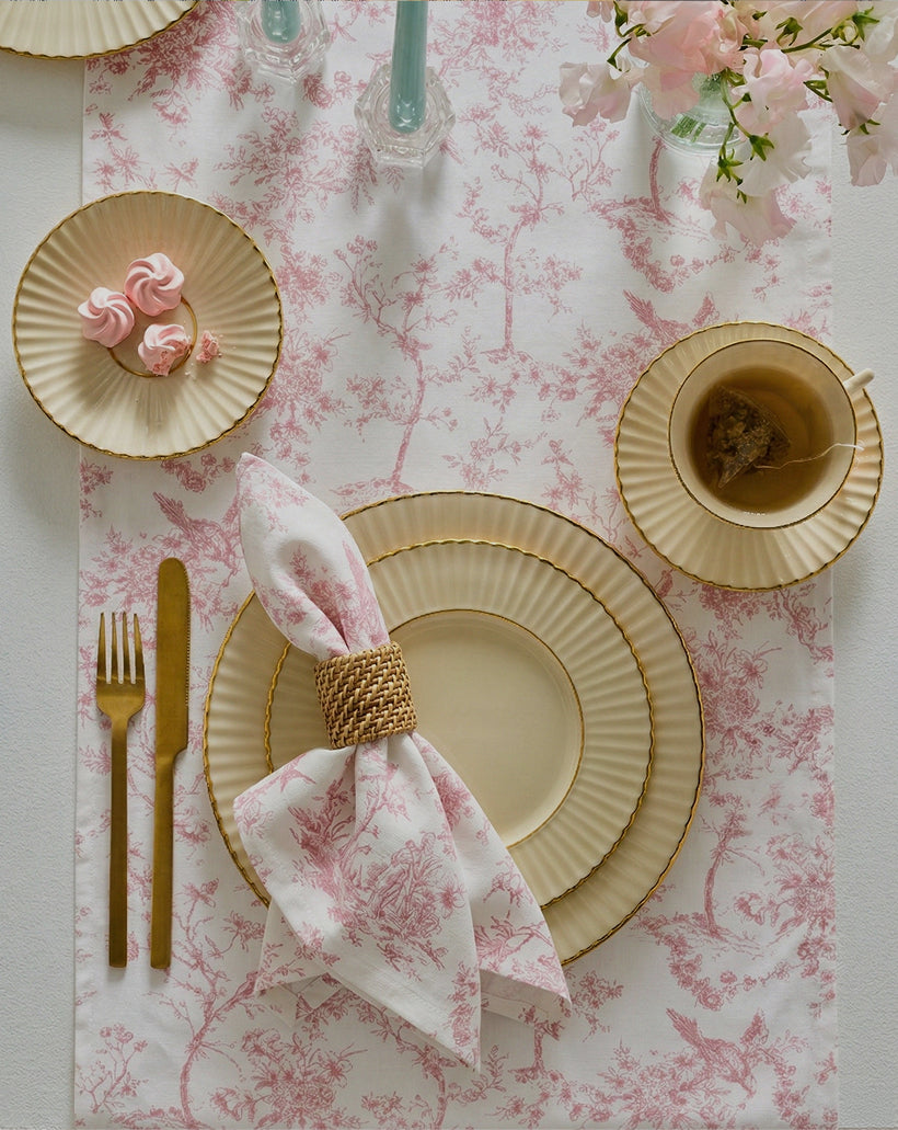Elegant table setting with cream and gold-rimmed plates, gold flatware, a teacup with tea bag, pink meringues on a side plate, a floral napkin in a woven ring, and pink flowers in a vase on a toile table runner.