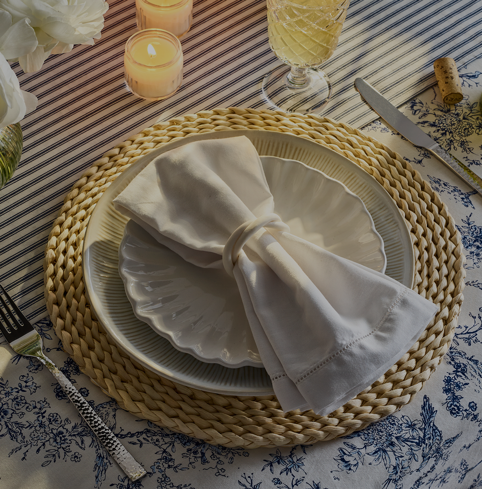 A place setting with a woven placemat, two stacked white plates, a folded white napkin in a napkin ring, silverware, a glass with a drink, candles, and a floral tablecloth.