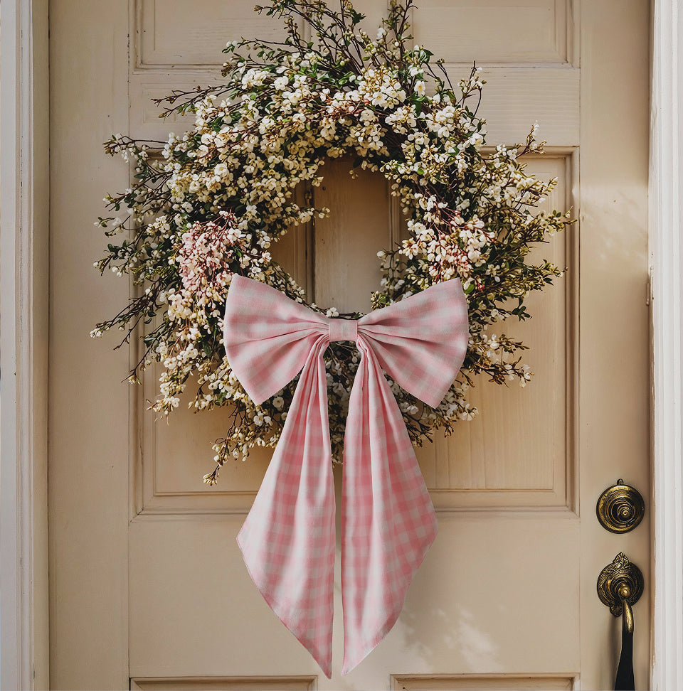 A spring wreath made of small white and pink flowers hangs on a cream-colored door, decorated with a large pink plaid bow at the bottom.