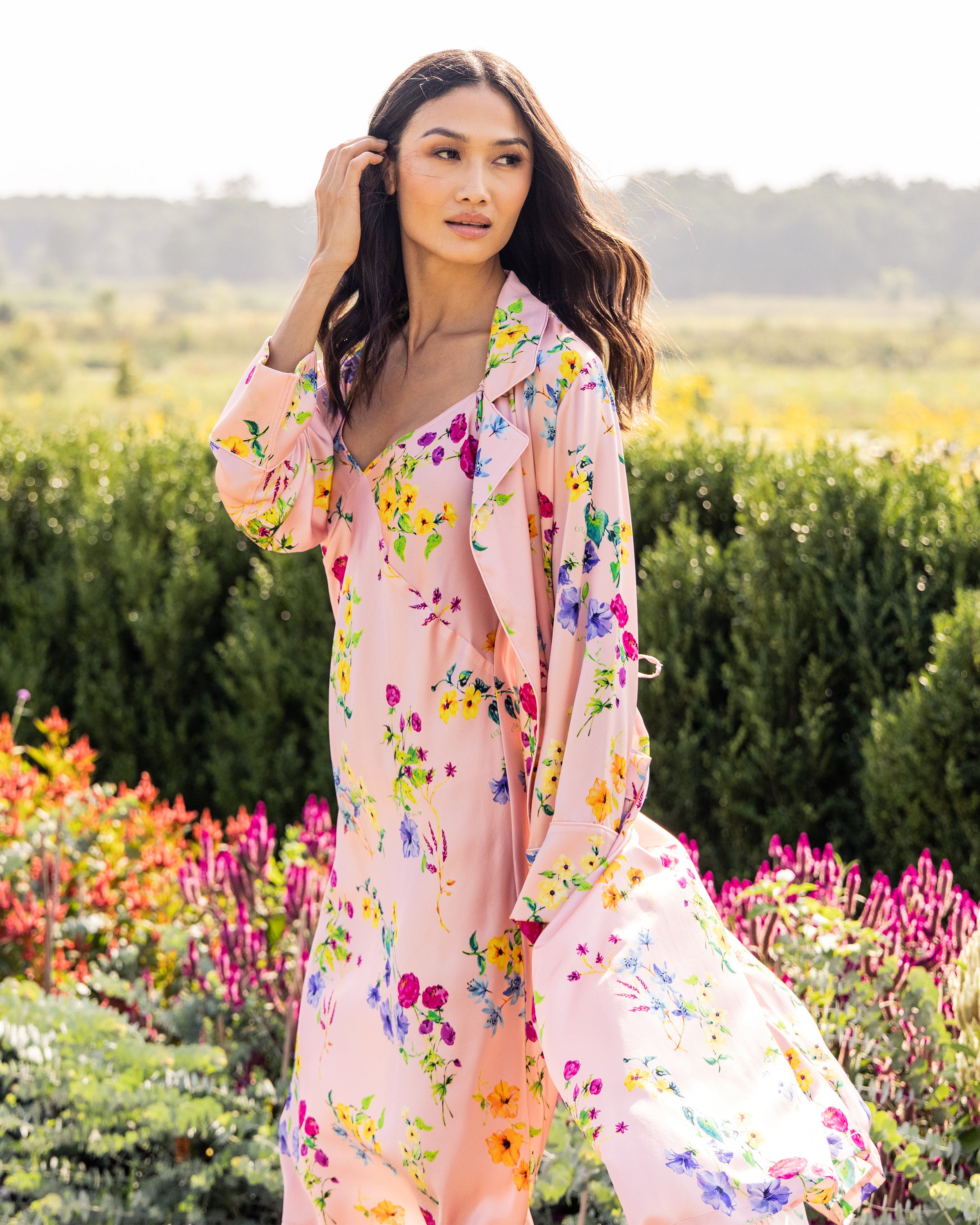 In a botanical garden, a person with long wavy hair holds it back, wearing Petite Plumes Womens Silk Robe in Blush Brilliant Botanical. Surrounded by colorful flowers, they stand against a backdrop of greenery and clear skies.