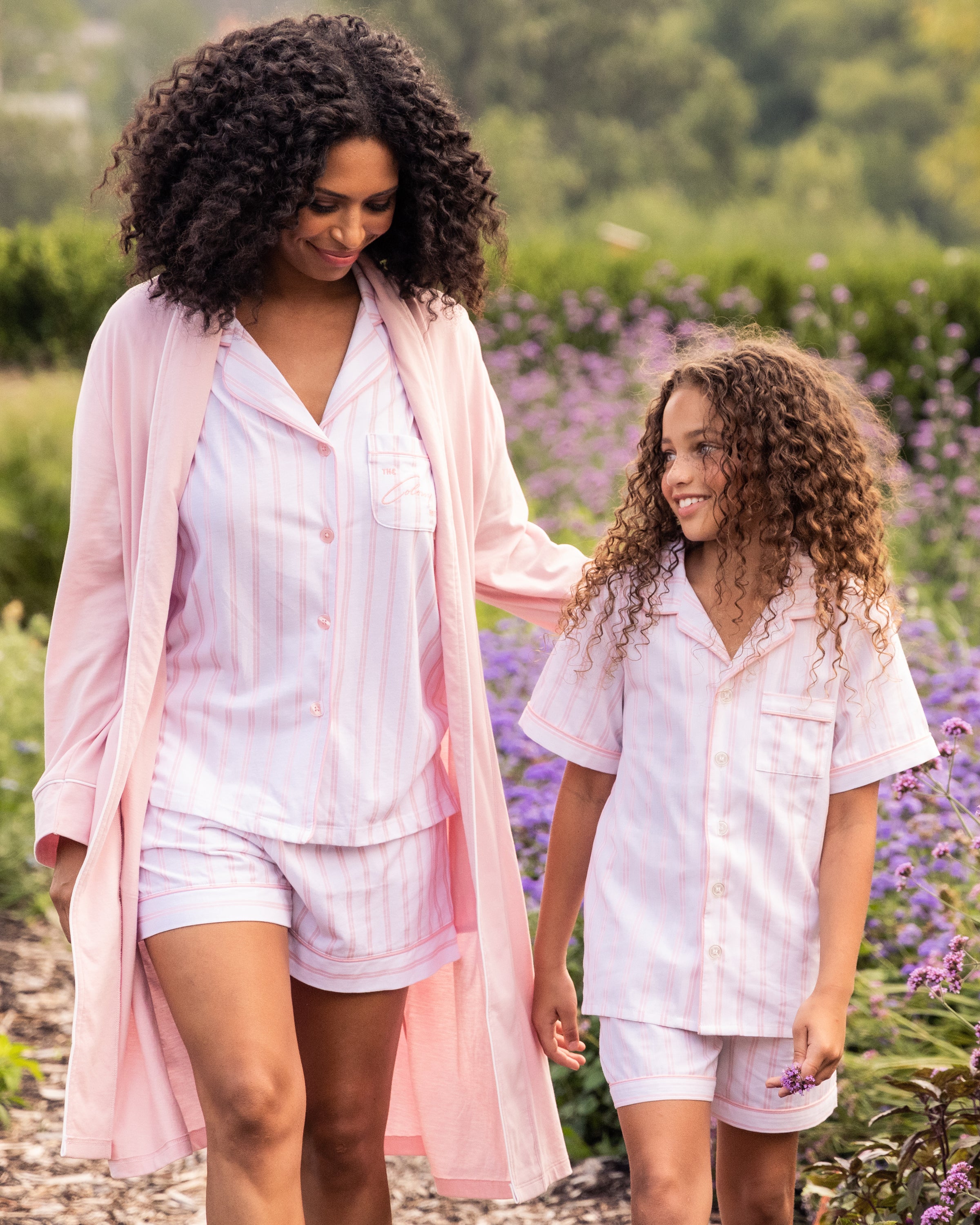 A woman and a young girl, both in pink and white striped pajamas made from luxurious Peruvian Pima cotton, walk outdoors. The woman wears the Women’s Pima Robe in Pink by Petite Plume over her breathable ensemble. They smile amid lush greenery and purple flowers.