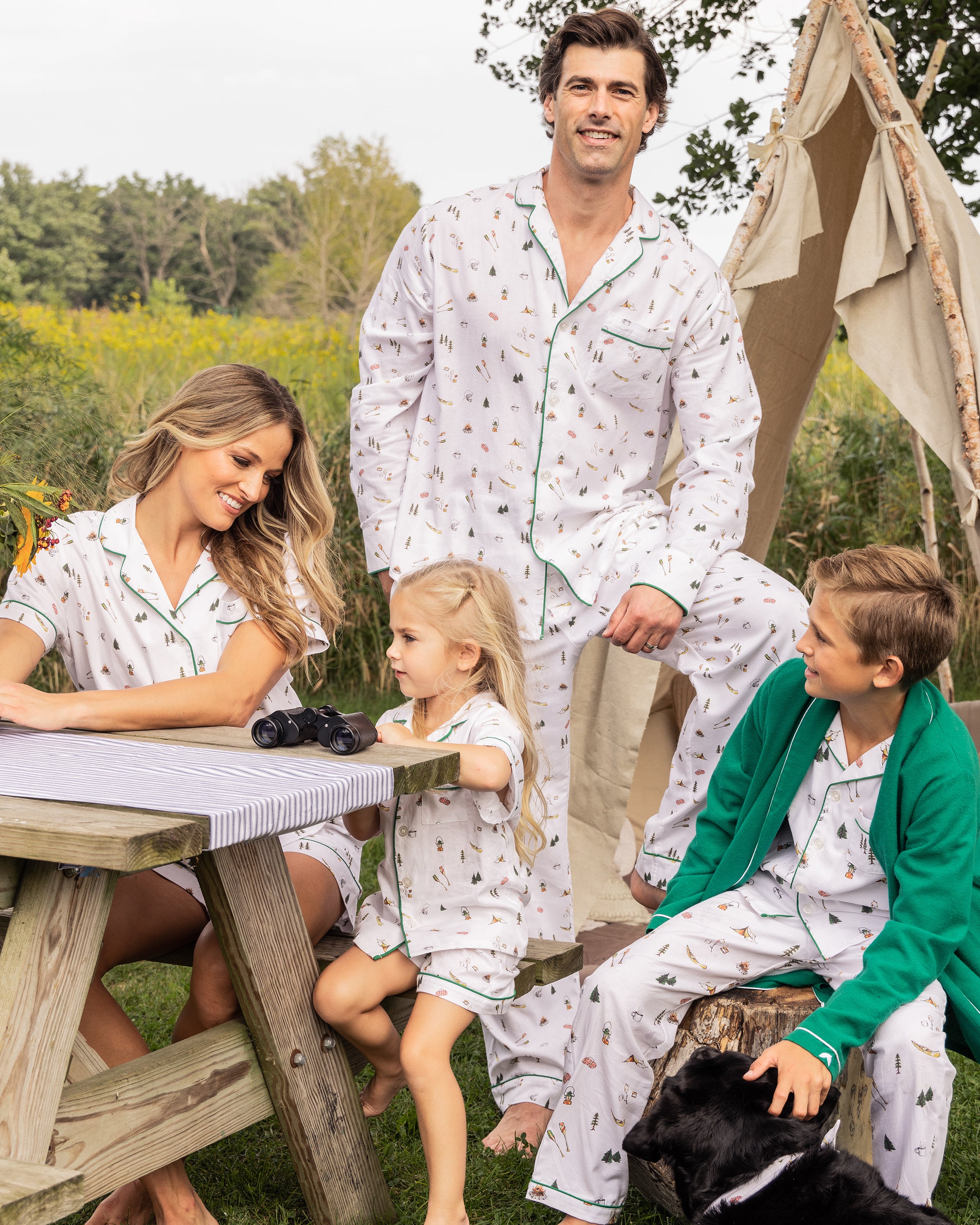 A family in matching Petite Plumes Mens Twill Pajama Set in The Great Outdoors with green trim gathers by a wooden picnic table. The woman and girl sit while the man stands, and the boy in a green sweater sits near a black dog, crafting an idyllic camping scene.