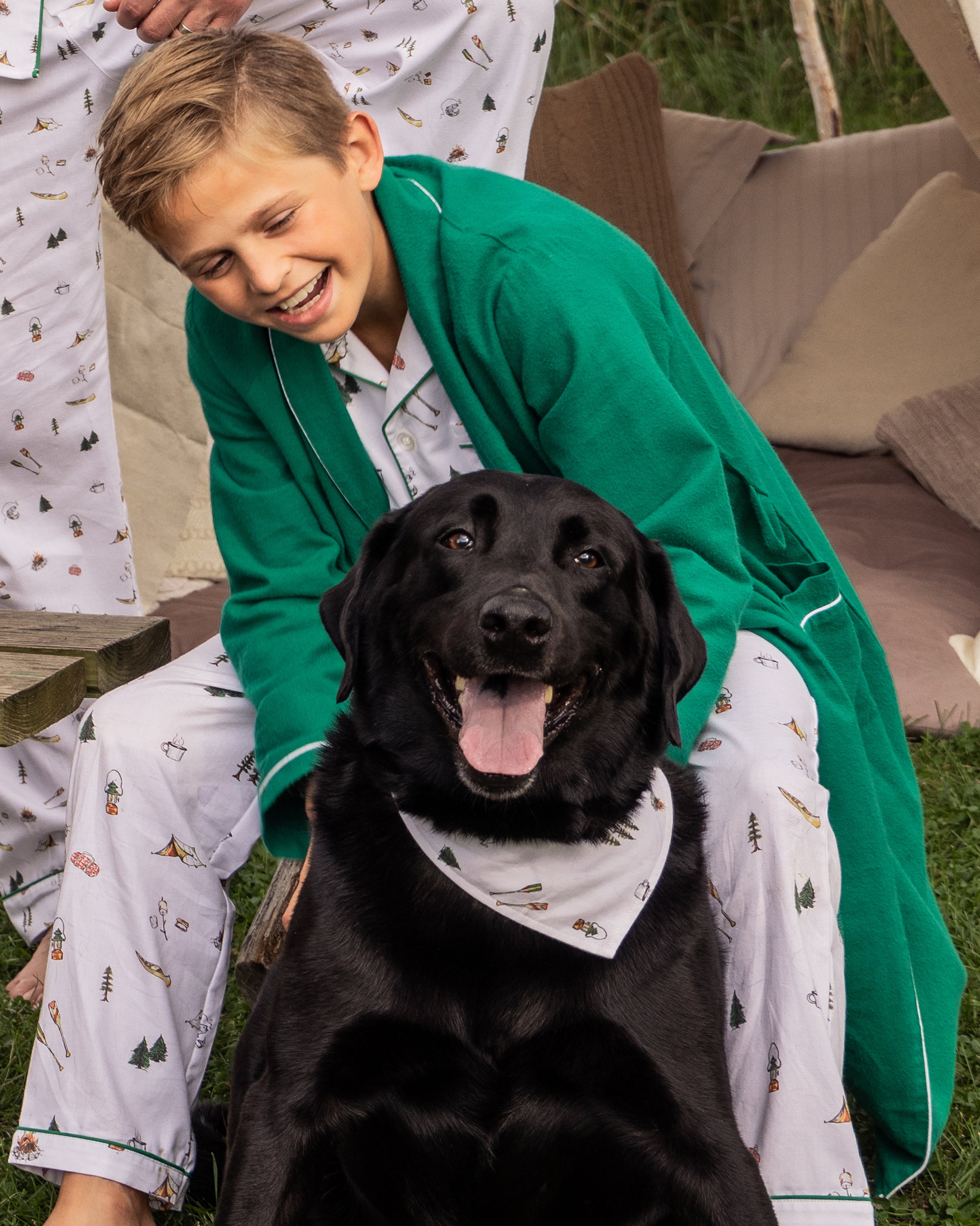 A young boy in a green robe and ski-patterned pajamas smiles behind a black Labrador sporting a matching The Great Outdoors bandana from Petite Plume. Both appear happy outdoors, surrounded by pillows and cushions.