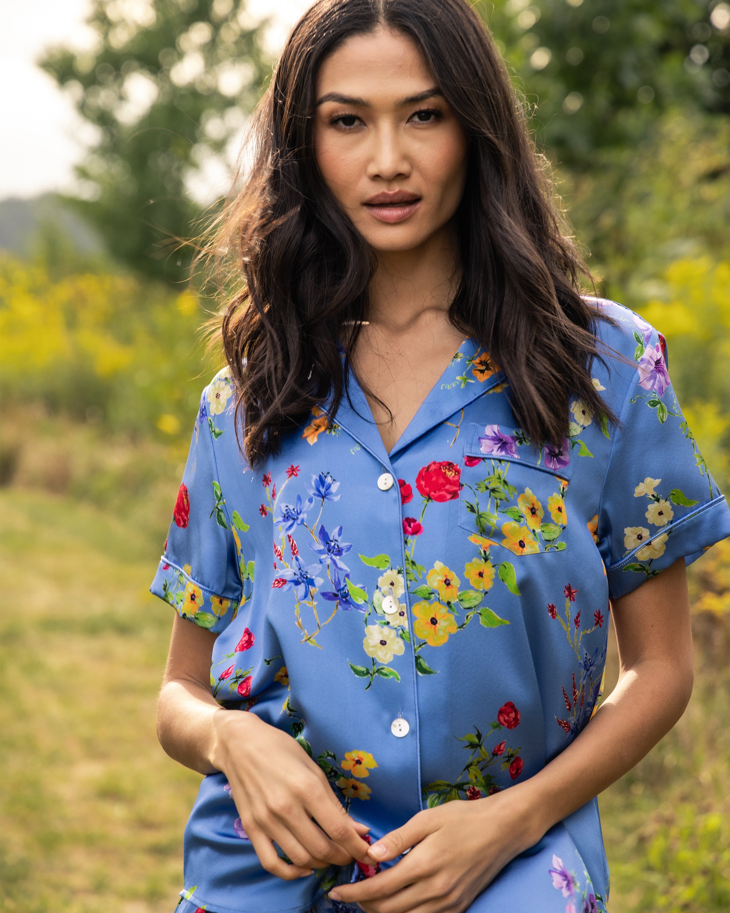 A woman with long dark hair stands outdoors in a grassy area, trees and yellow flowers behind her beneath a partly cloudy sky, wearing Petite Plumes Womens Silk Pajama Short Set in Brilliant Botanical Azure.