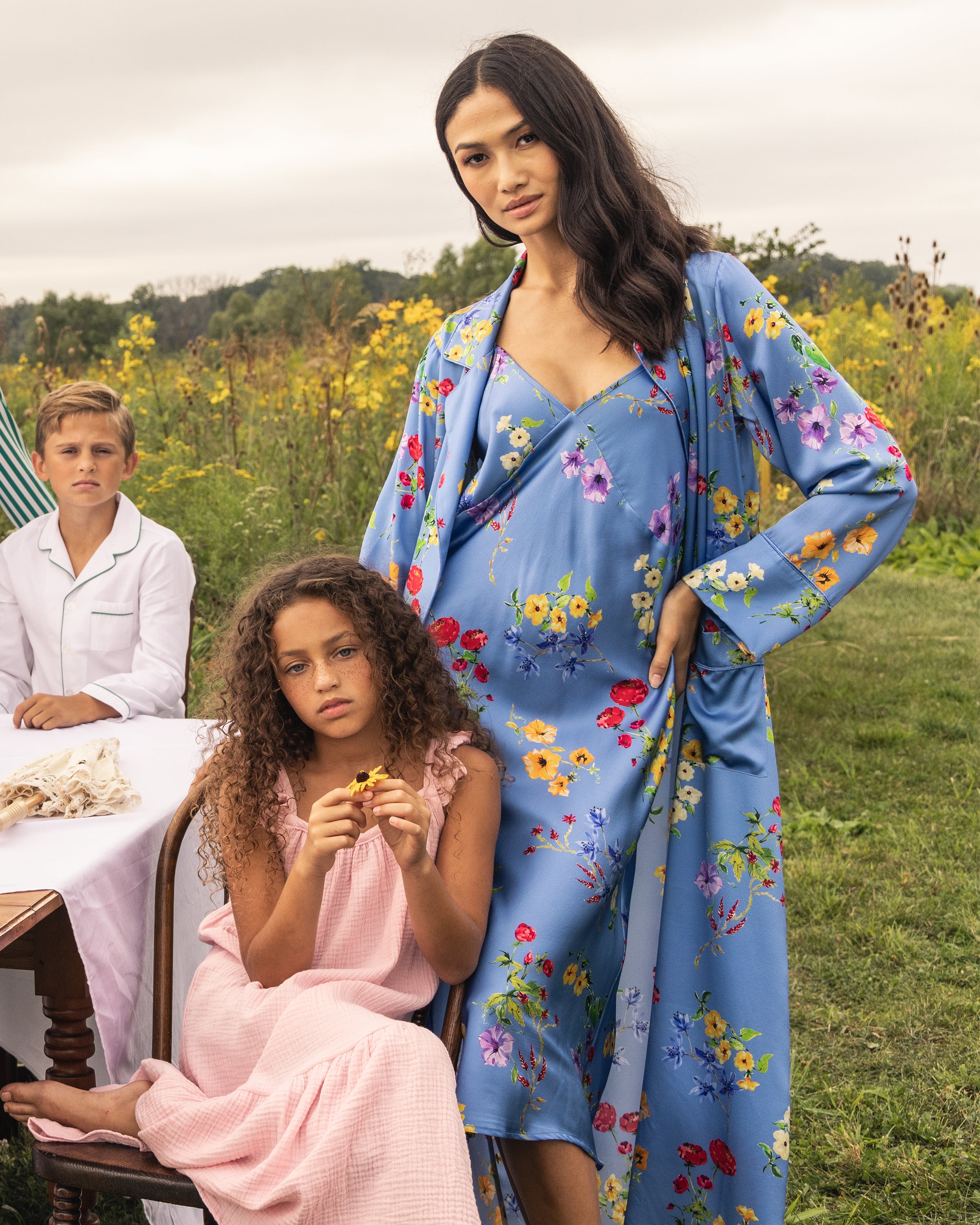 A woman in a Silk Cosette Nightgown in Azure Brilliant Botanical by Petite Plume stands in a field, next to a girl on a bench in a pink print dress. A boy sits nearby amidst flowers and greenery under the cloudy sky, evoking serene luxury sleepwear dreams.