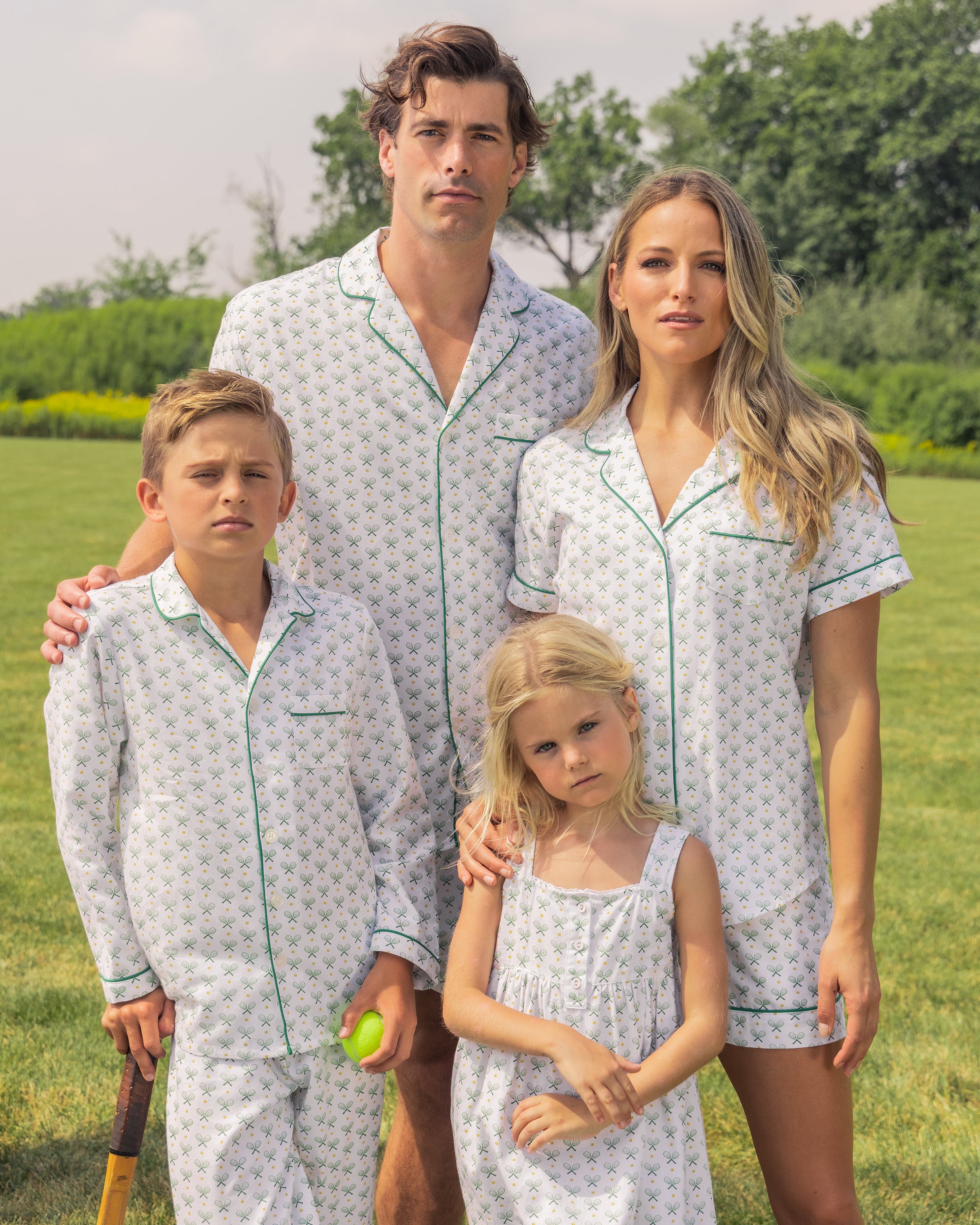 A family of four stands on a grassy field in matching Petite Plume tennis print pajamas, including the Girls Twill Charlotte Nightgown in Match Point. The father and son hold croquet mallets as trees dot the background under a partly cloudy sky.