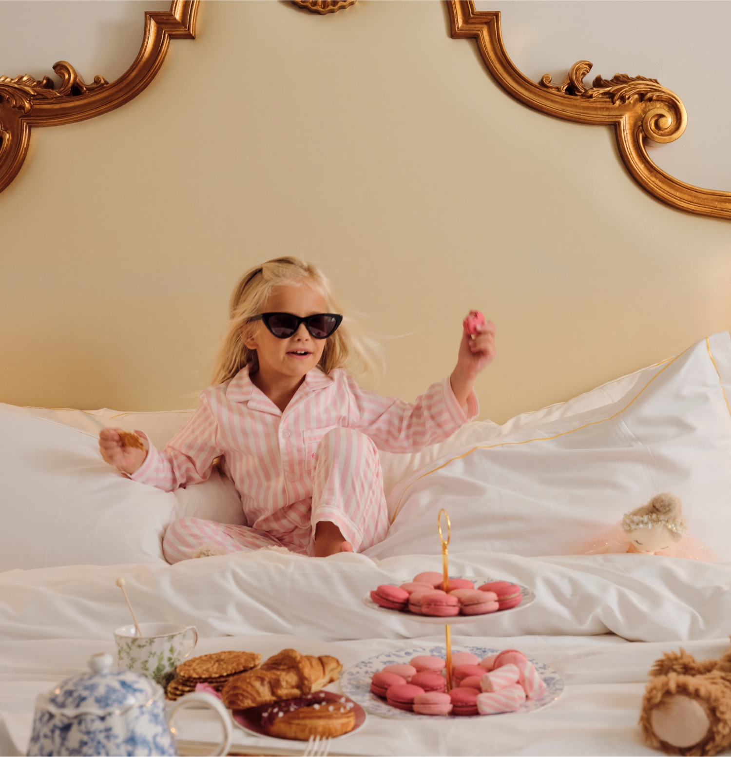 A young girl in pink striped pajamas and sunglasses sits on a bed with a gold ornate headboard, smiling and holding a macaron. A spread of pastries, tea, and macarons is arranged on the bed in front of her.