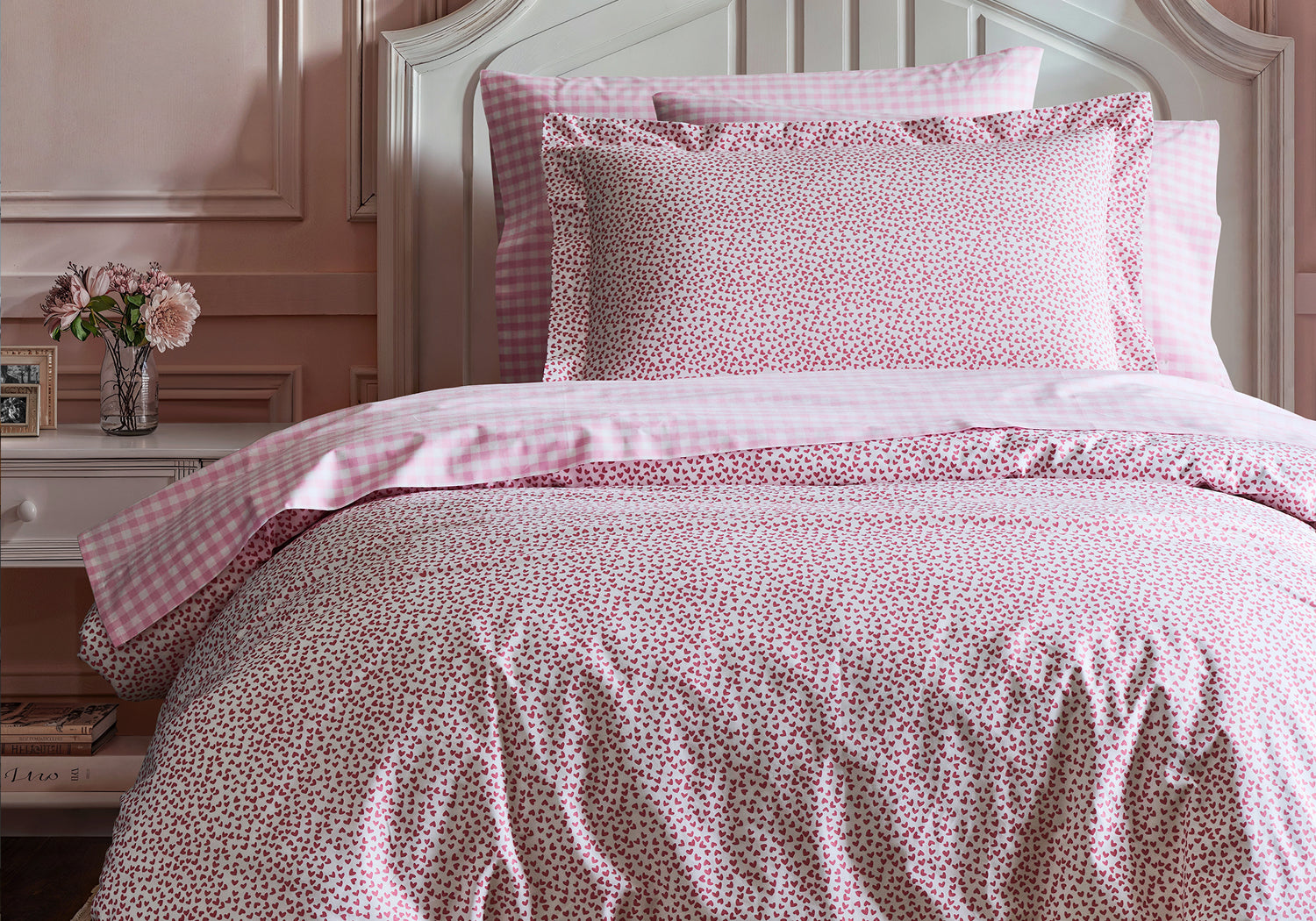 A neatly made bed with pink floral and gingham bedding, set against a light pink wall. A white nightstand with a vase of flowers, a photo frame, and books is beside the bed.