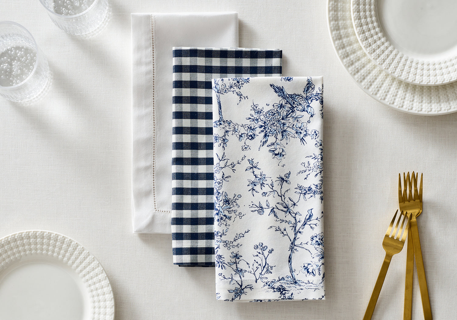 Three neatly folded cloth napkins—one white, one navy checkered, and one with blue floral prints—are arranged side by side on a set table with white plates, clear glasses, and gold utensils.
