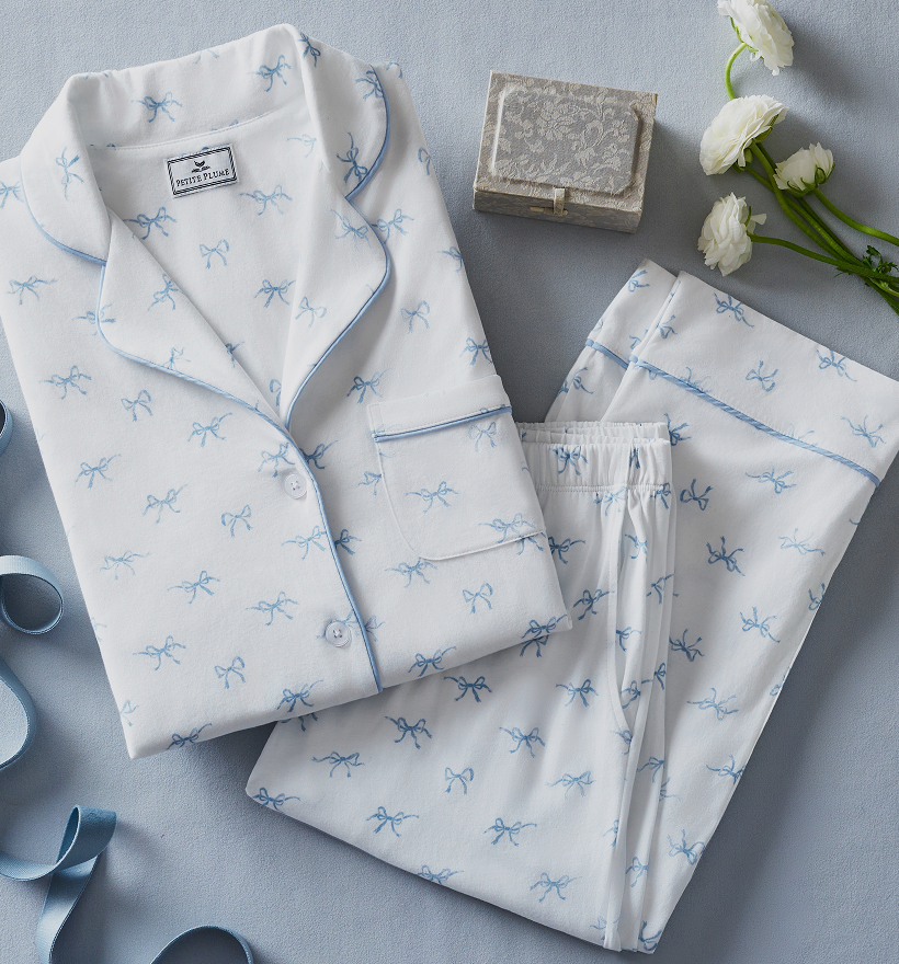 A set of white pajamas with blue bow patterns, neatly folded next to a small lace-covered box and three white flowers on a light blue surface. A blue ribbon is also partially visible.