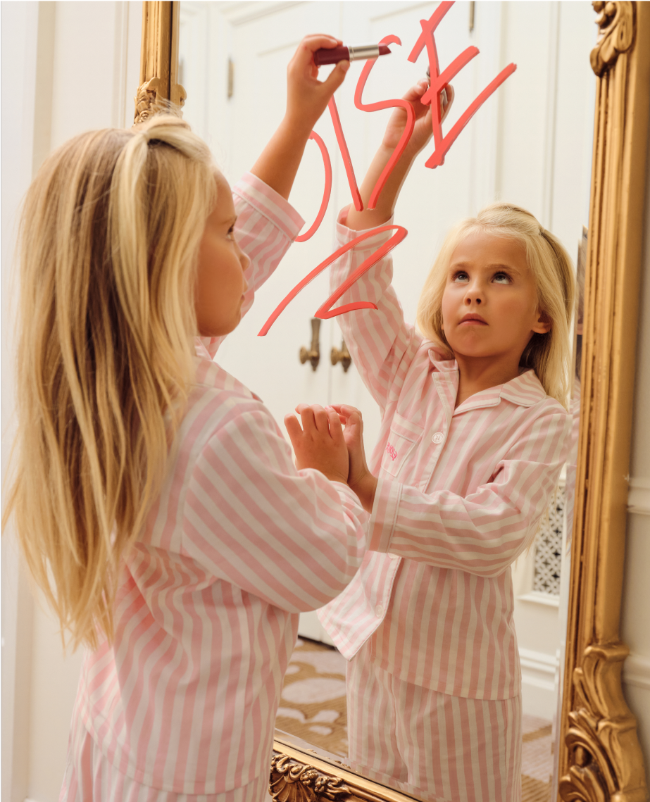 A young girl in pink and white striped pajamas writes YES on a mirror with red lipstick, her reflection visible in the ornate gold-framed mirror.