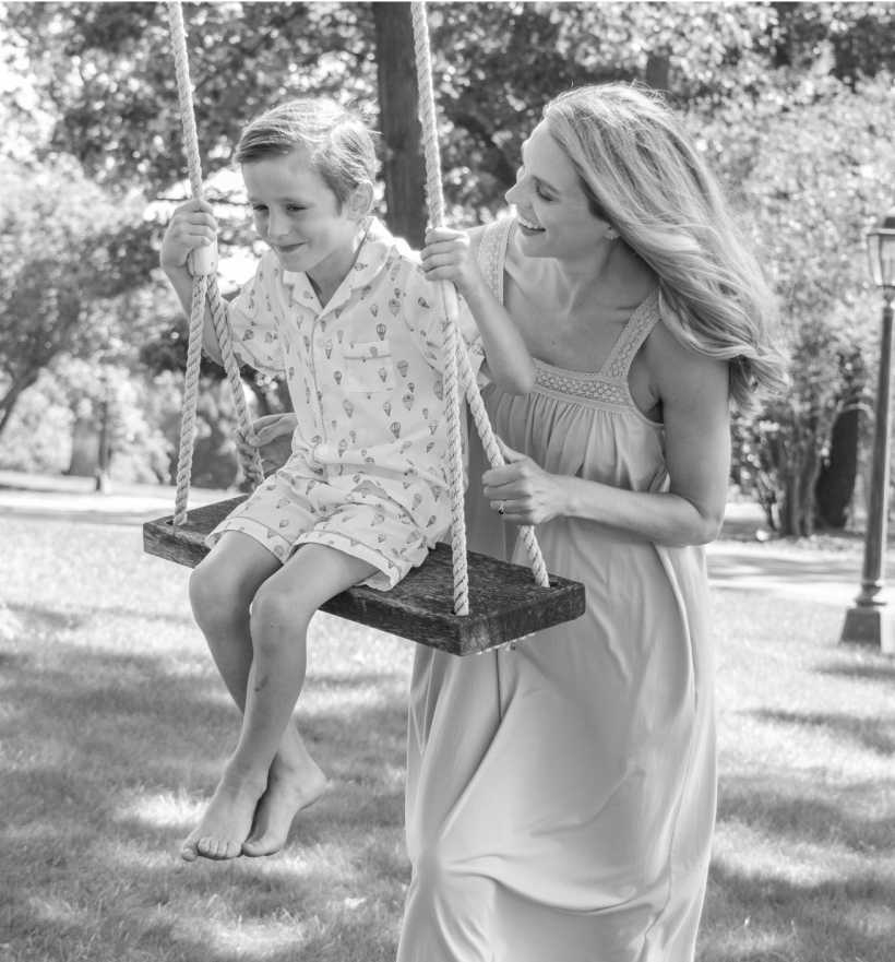 Black and White photo of a Mother and Child outside. The Mother is pushing the child on swing under a tree
